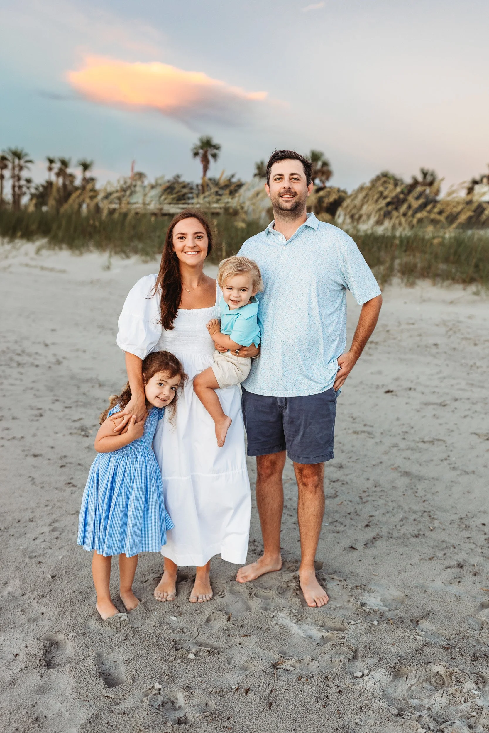 family on th beach at sunrise in front of a dune holding two small toddlers who are laughing