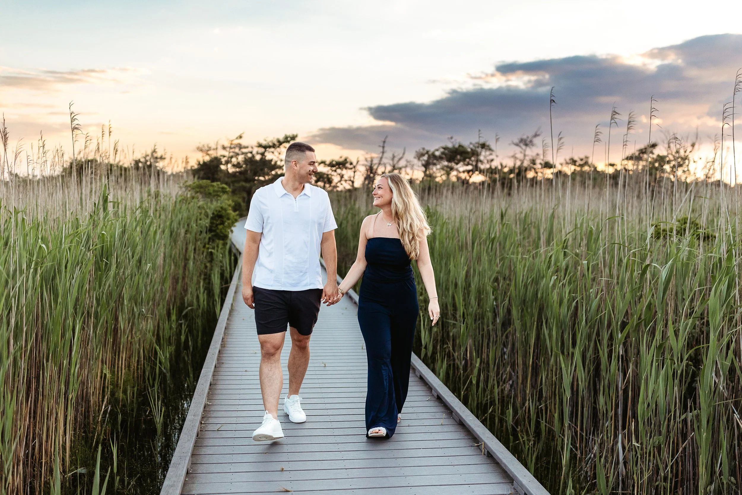 A couple walking hand in hand on a wooden boardwalk through tall green reeds at sunset, smiling at each other.