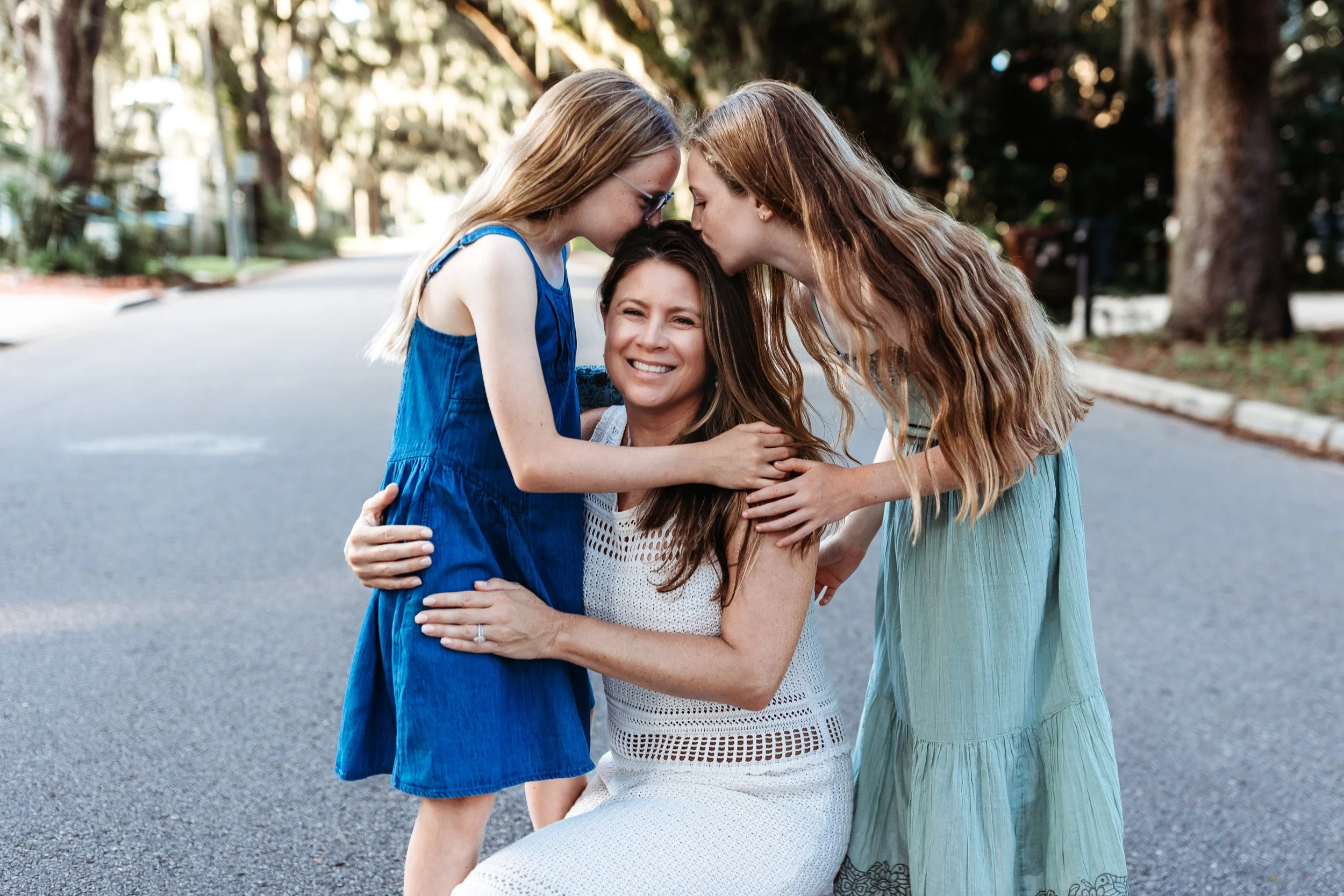twin daughters kissing mom who is kneeling down in the street with moss trees behind them
