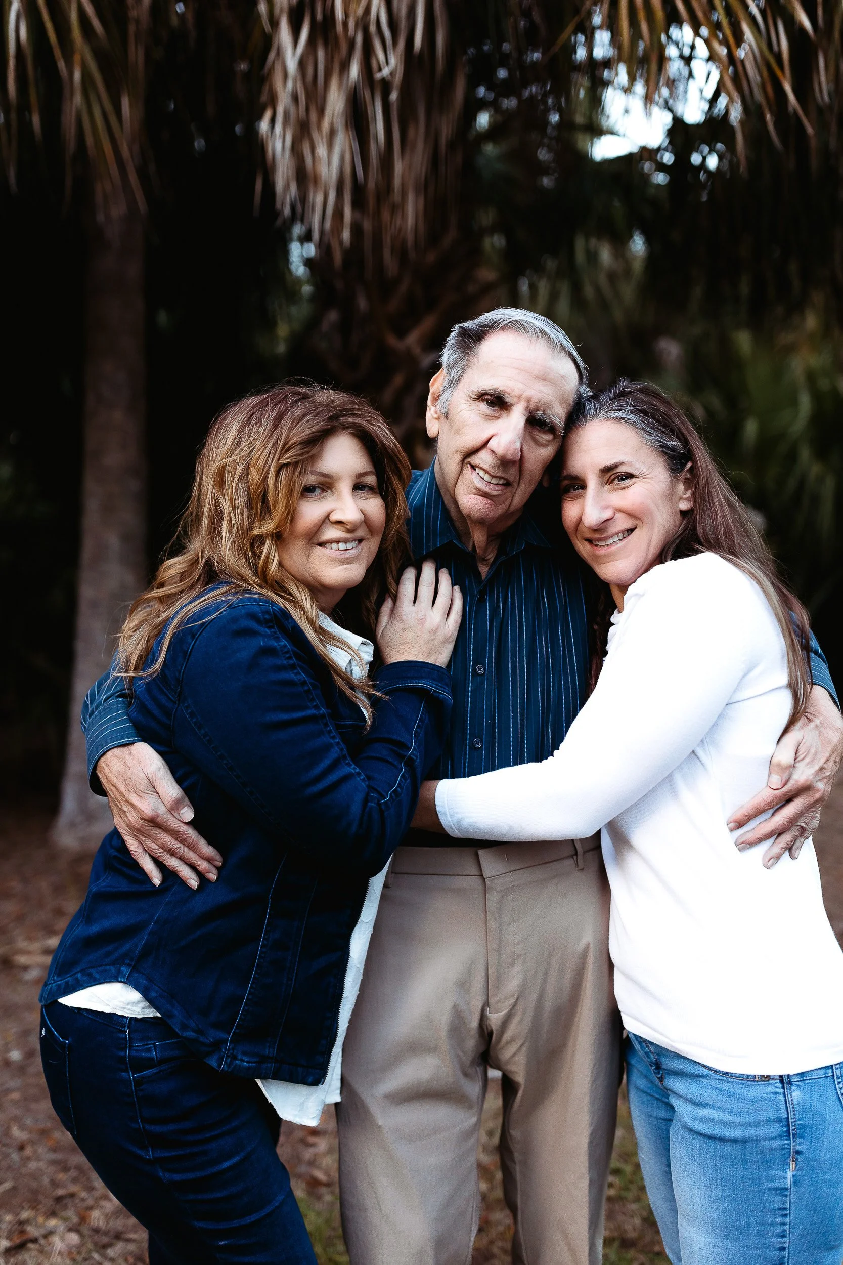adult daughters embrace their elderly dad with palms and oak trees behind them at twenty mile park