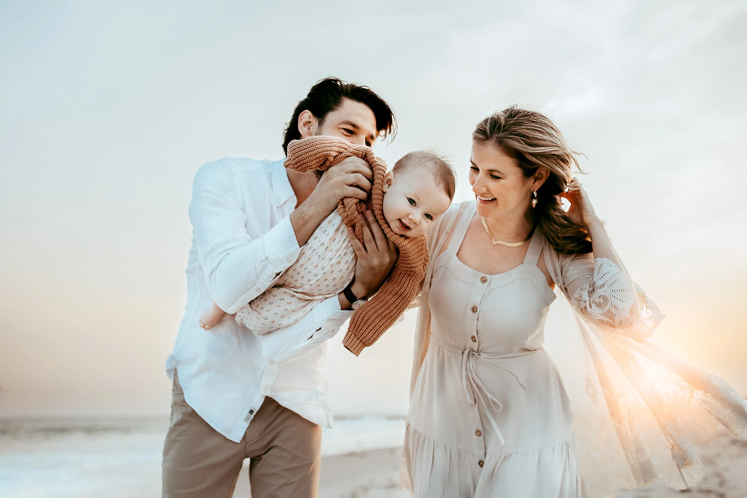 dad flying baby girl in air at sunset for Ponte Vedra Beach family photos at the beach and moms lace shawl is blowing in the wind with the sun shining through .  they are both laughing 