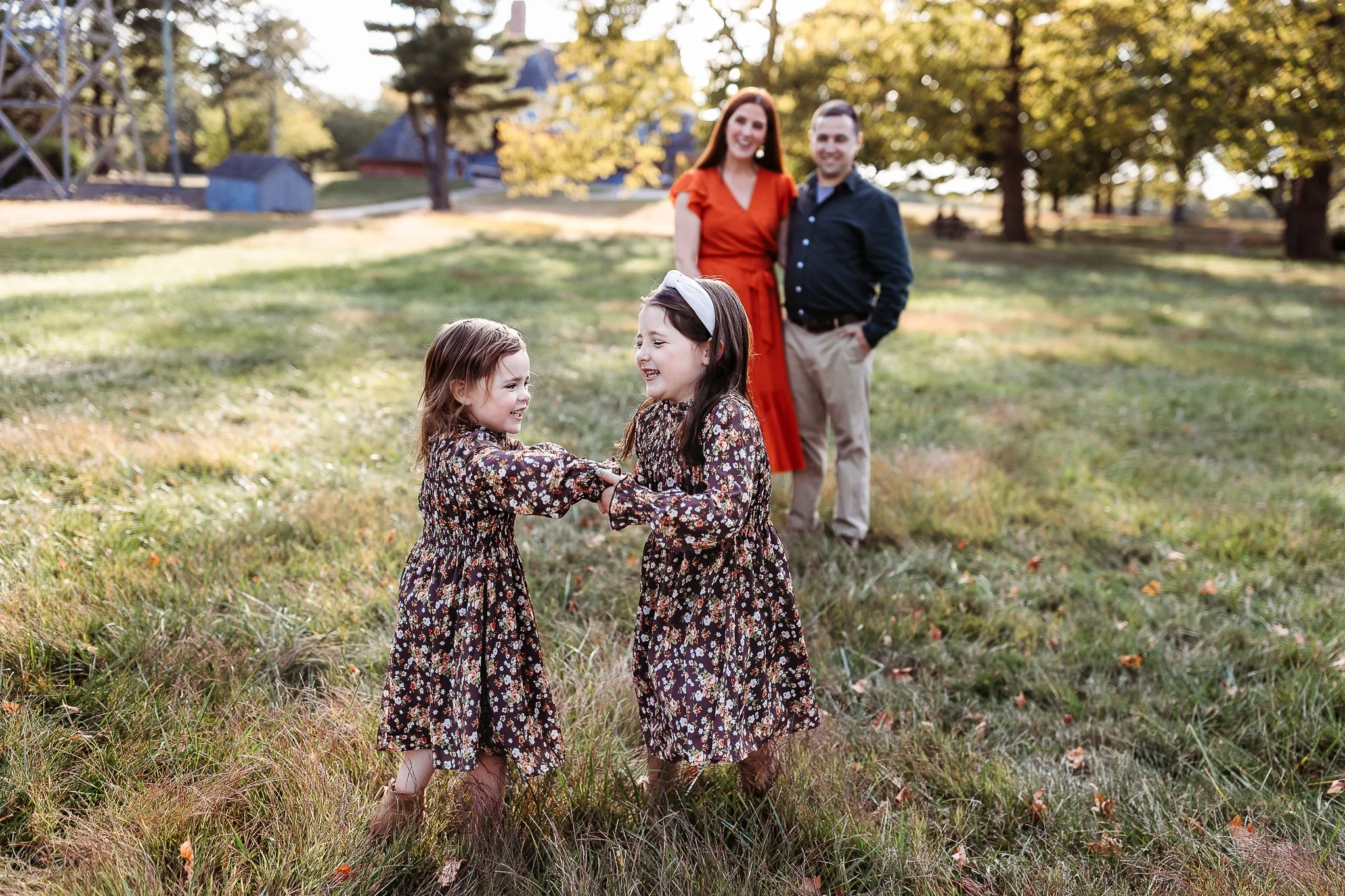 sisters in matching brown floral dresses on a farm.  they are holding hands and twirling .  parents in the background embraced and looking at them and smiling