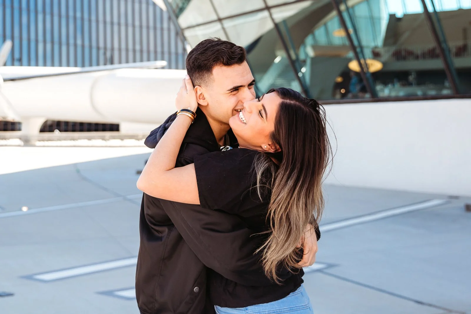 A young couple embraces and smiles happily on a yacht deck, with a modern glass building in the background.