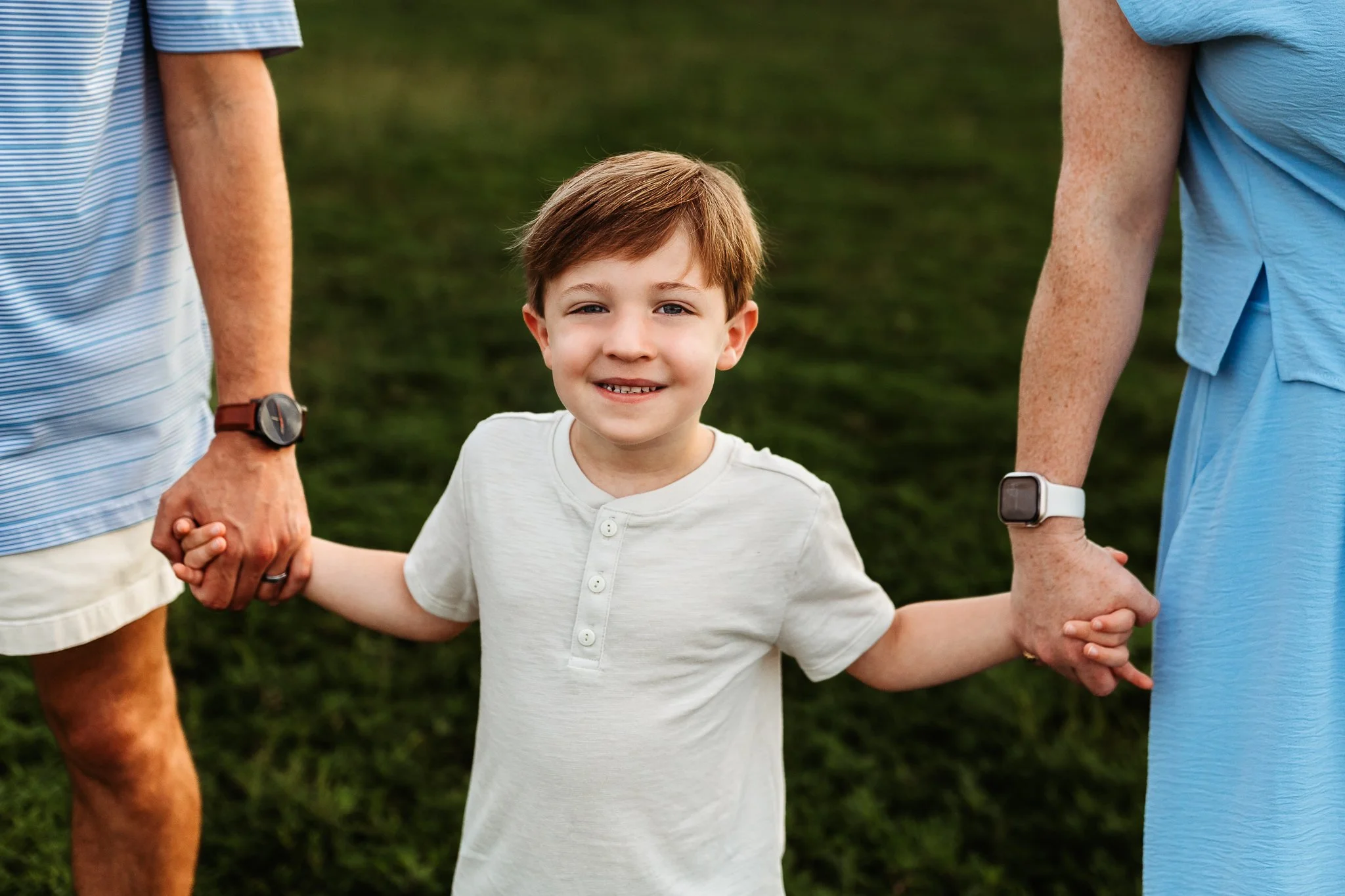 close up of a boy holding parents'hands while walking on a green lawn for family pictures