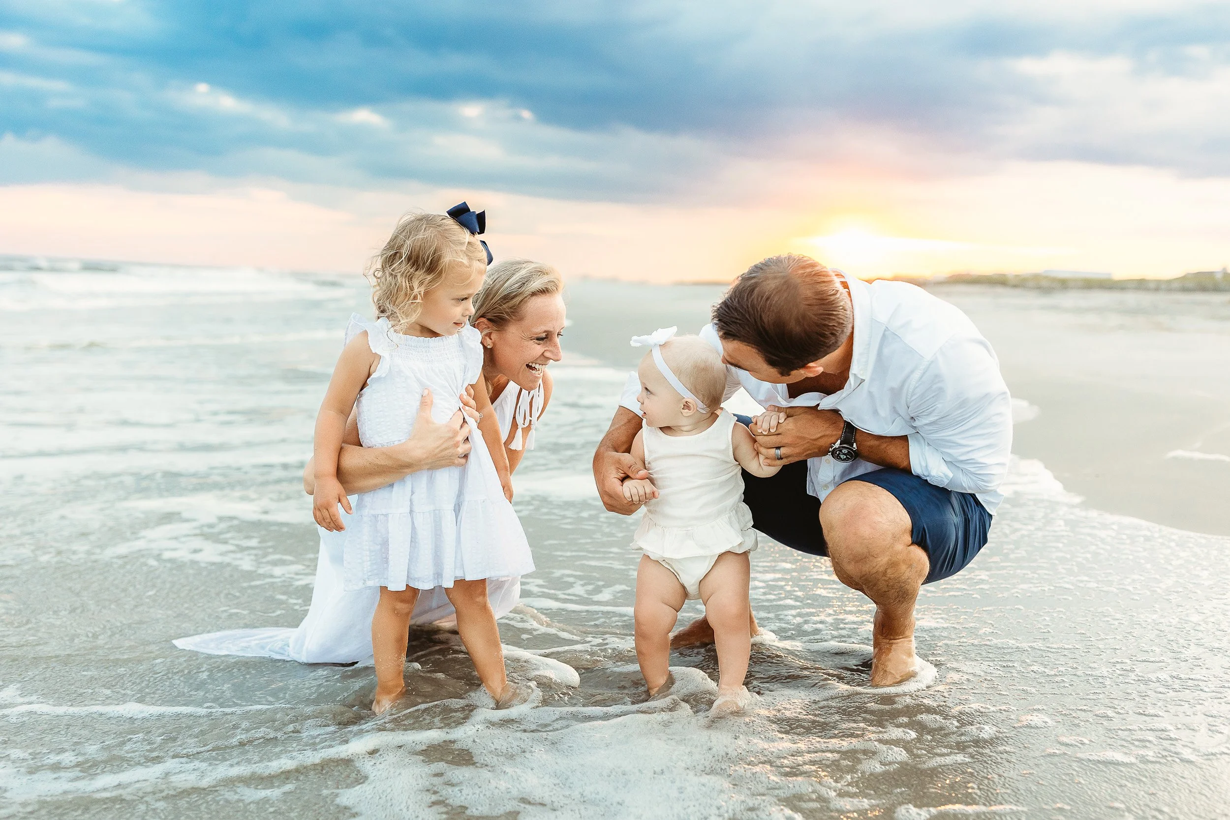 family crouched in the ocean with little toddler girls and they are laughing and the sunset is behind them