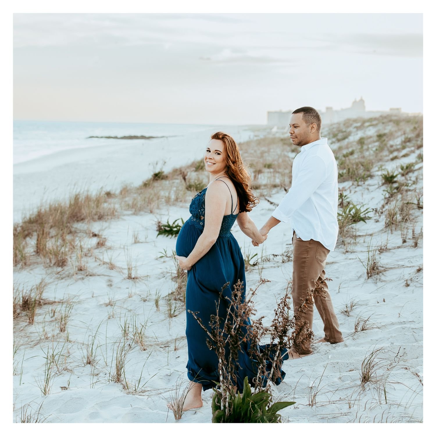 A pregnant woman in a blue dress and a man holding hands on a sandy beach with dunes and sparse vegetation, facing away from the ocean, during daylight.