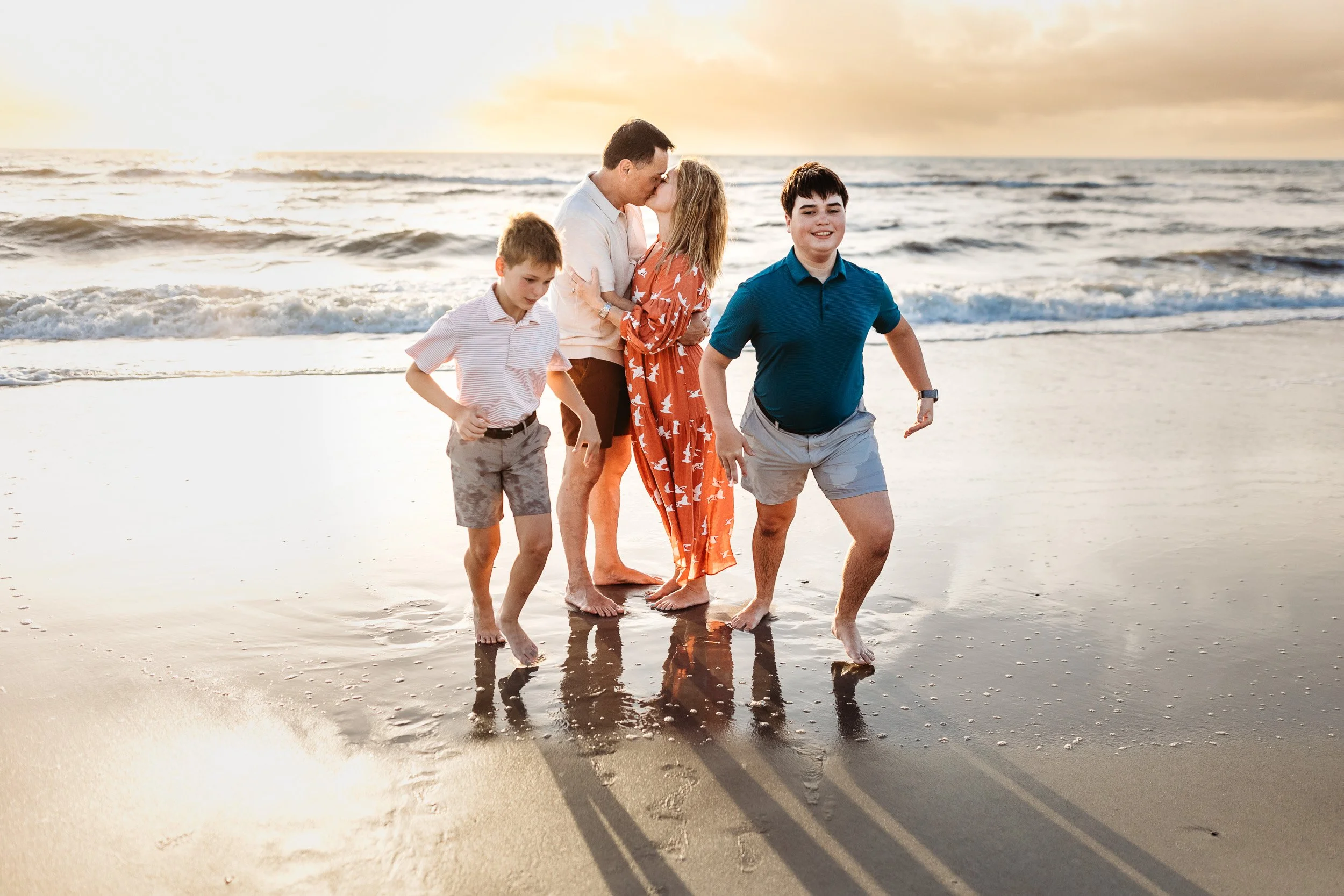mother and father kissing on th beach at sunrise while the sons run away from them laughing