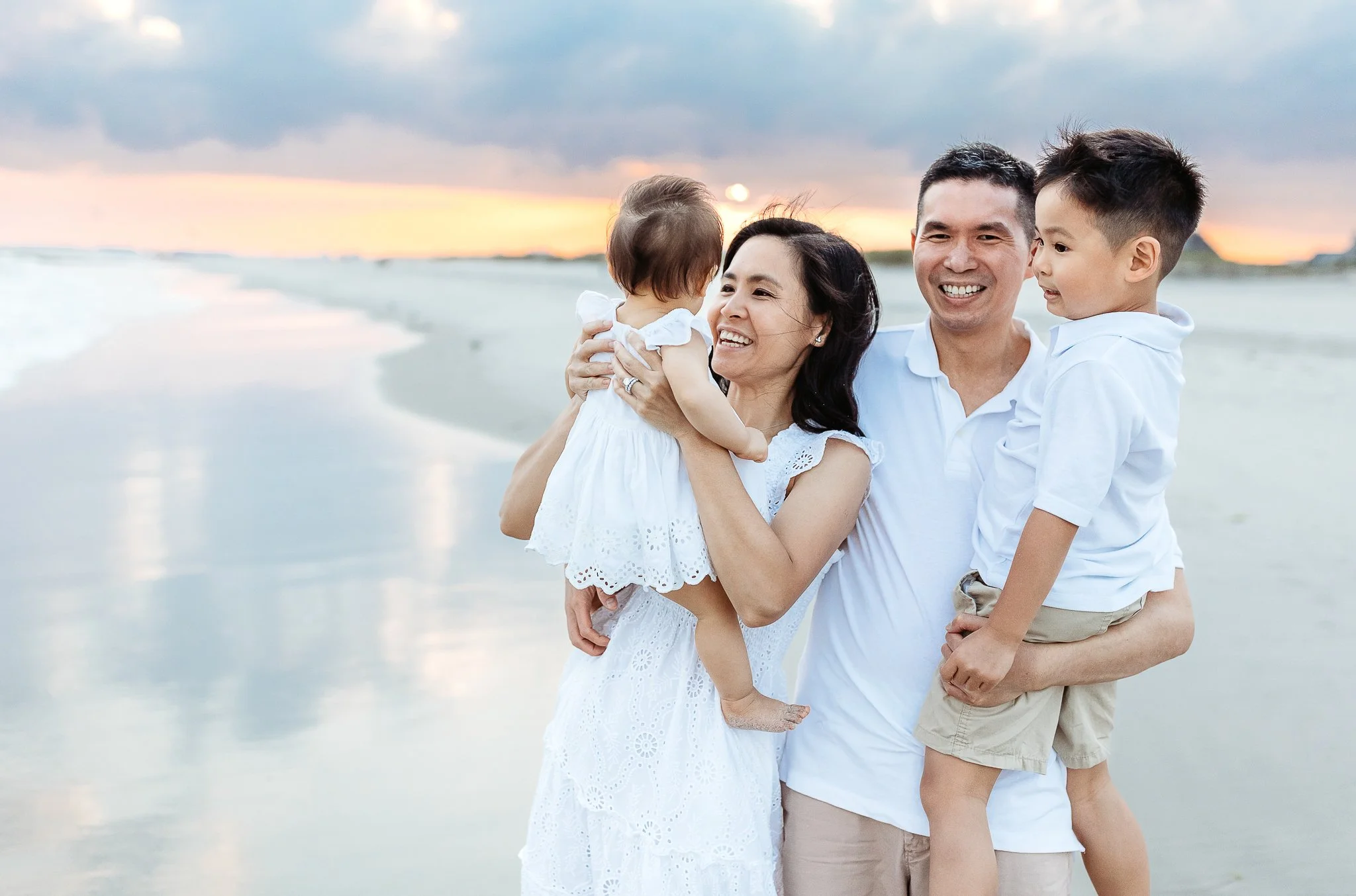 family standing at ocean's edge lifting kids while embracing and they are in all white and laughing