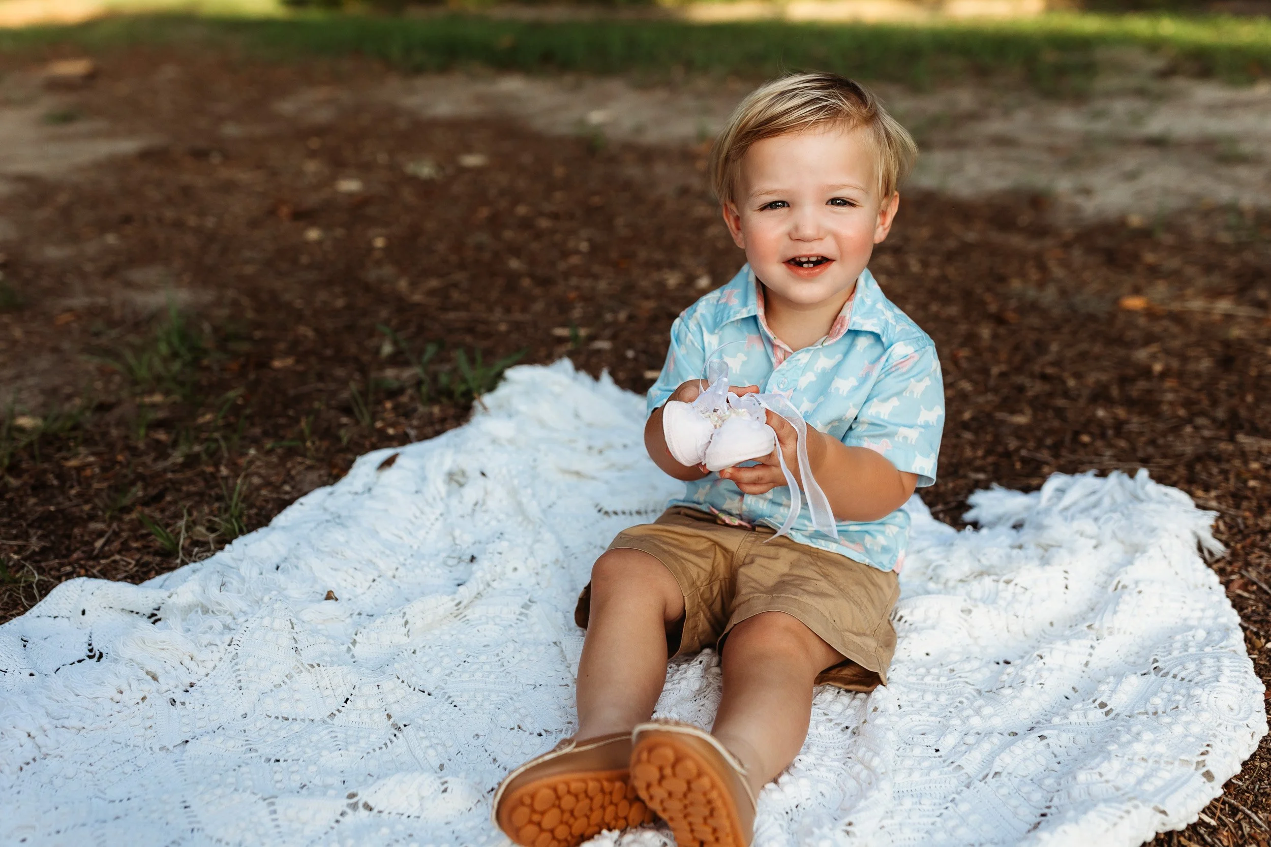 toddler boy holding pink baby shoes while seated on a crochet blanket in a park at sunset for maternity family photos