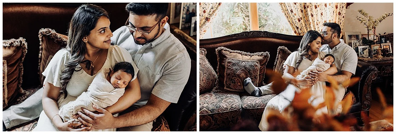 husband and wife seated on a wood couch in front of a window holding a baby and looking at each other