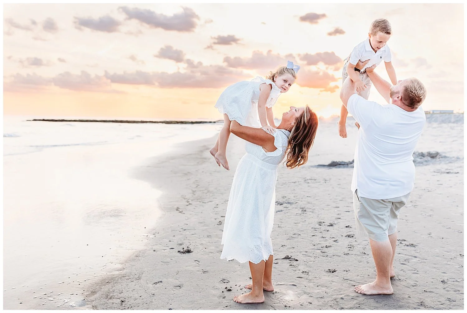 mom and dad lifting children into the air and laughing at the beach ocean behind them pink and orange cloudy sunset sky all in white outfits