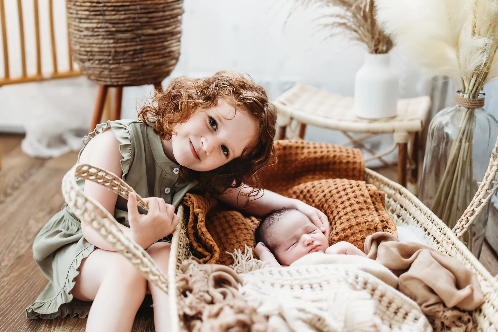 toddler sibling in olive romper with her baby sister in a. Moses basket with earth toned blankets during an in home newborn session in Jacksonville Beach
