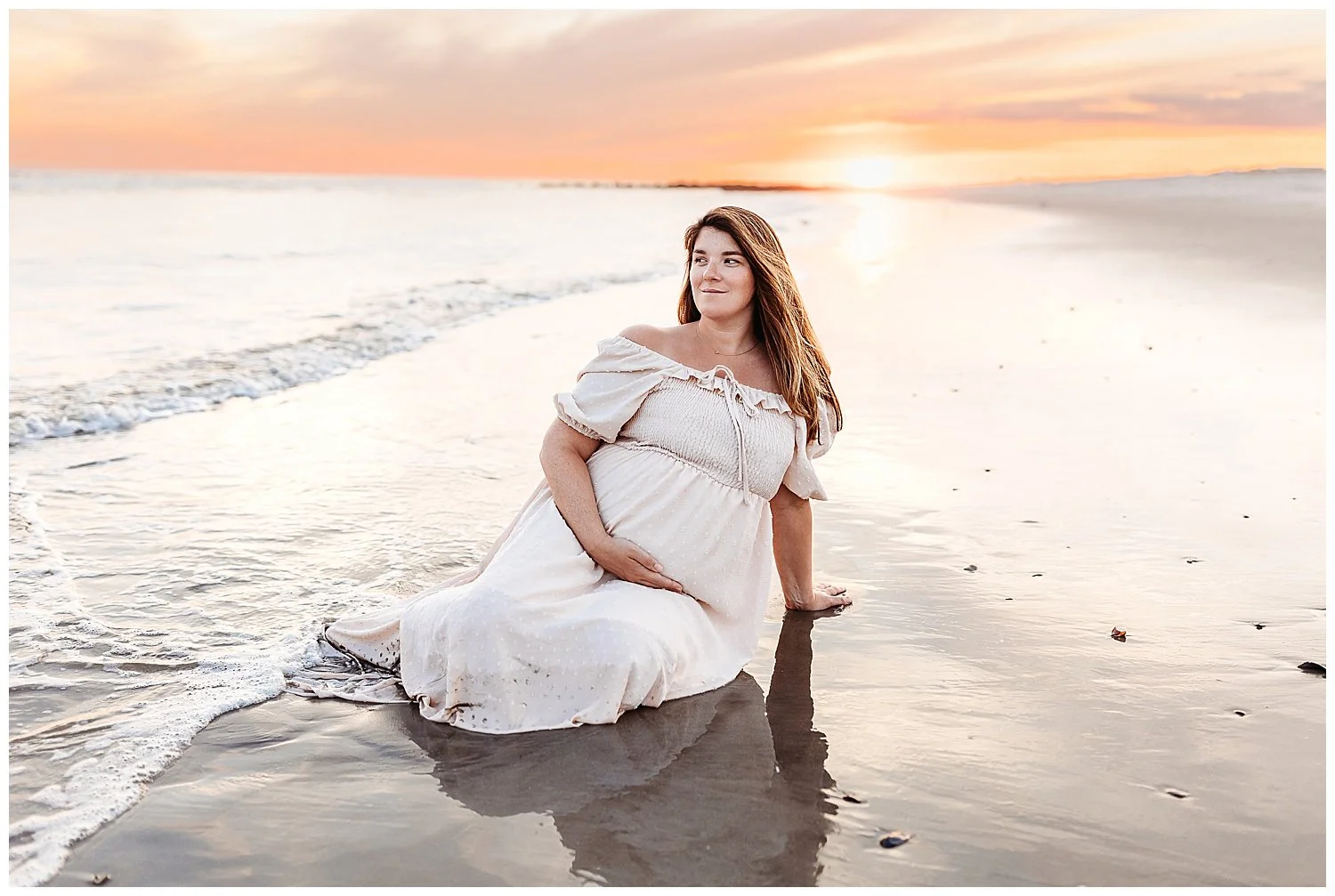 pregnant woman seated in the ocean at shore holding belly at sunset