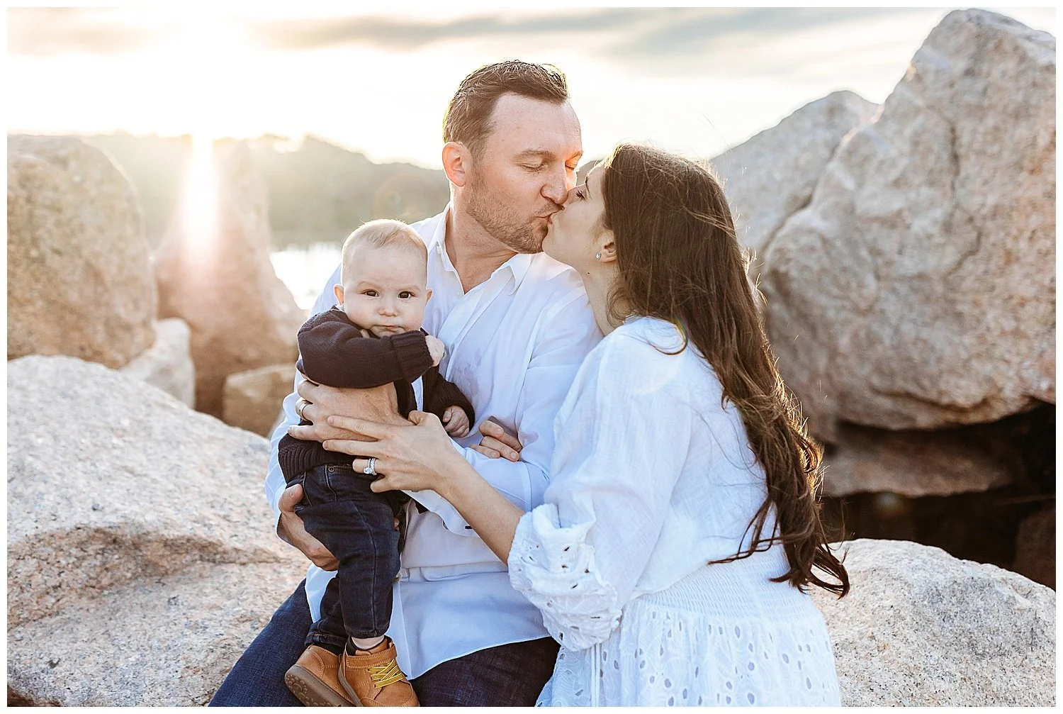 parents kissing while they hold a baby boy who is looking at the camera sunset behind them on a jetty beach