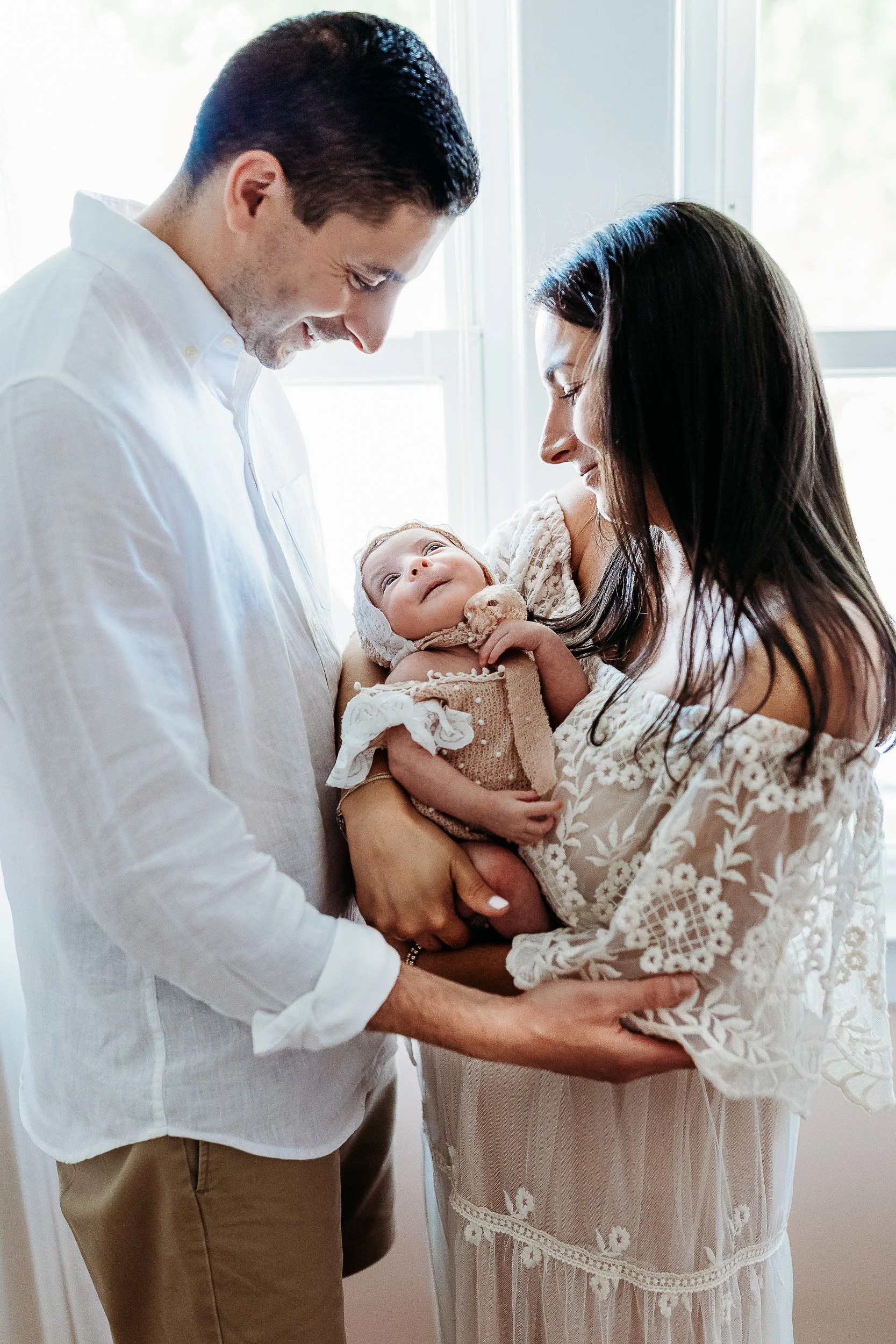 mother and child holding newborn baby girl in between them while standing in front of a window and mom is wearing a lace off the shoulder gown and they are taking photos in their home for a st augustine beach baby photo session