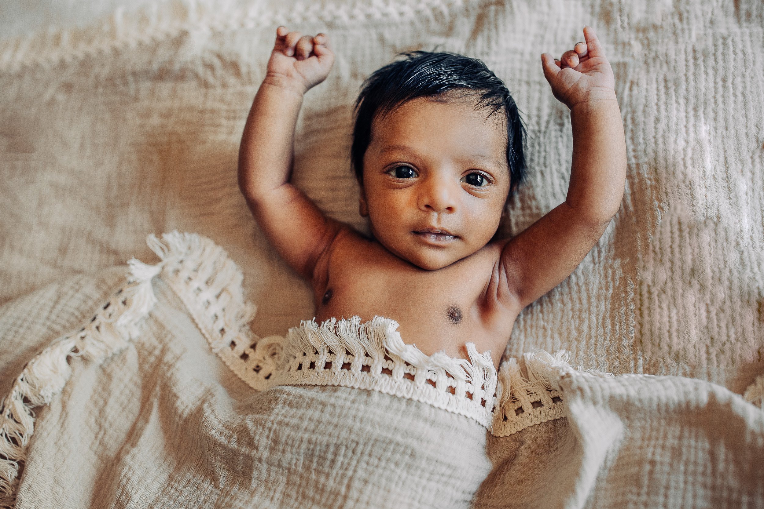 baby in muslin blanket with fringe stretched out and looking at the camera during in home newborn photos