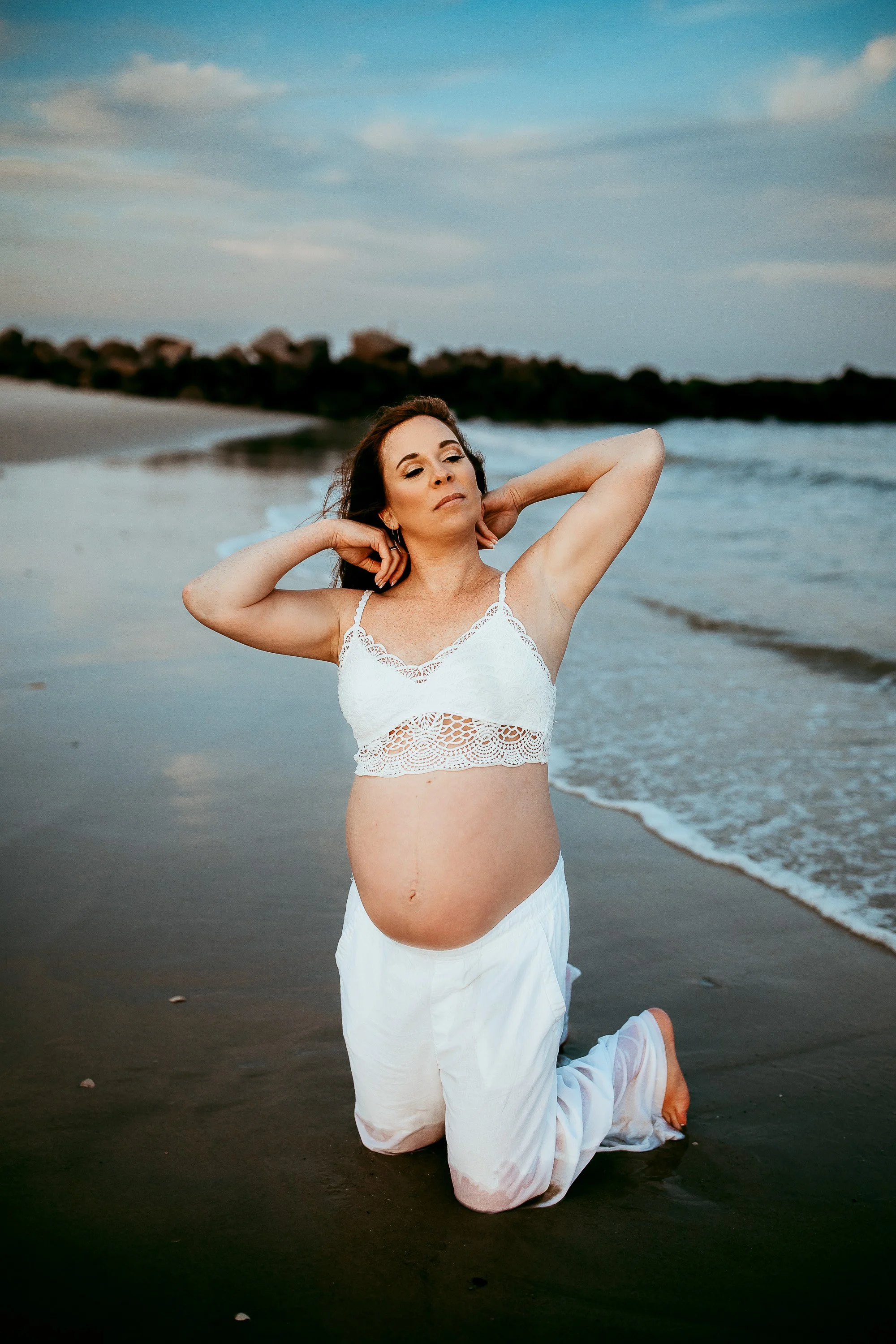 woman in a white bra and pants kneeling in the ocean during blue hour while holding her hair and looking up to the sky for maternity pictures on the beach in st augustine 