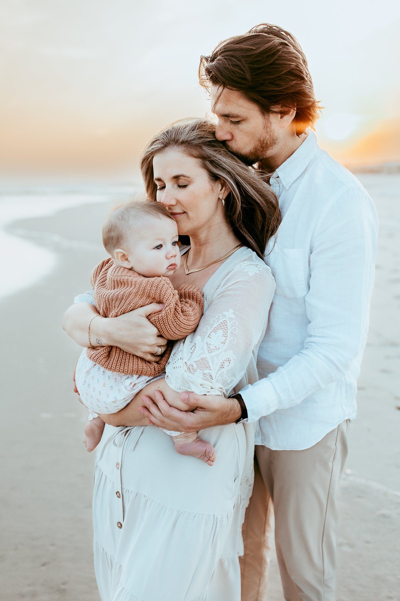 husband kissing wife while she holds baby at sunset