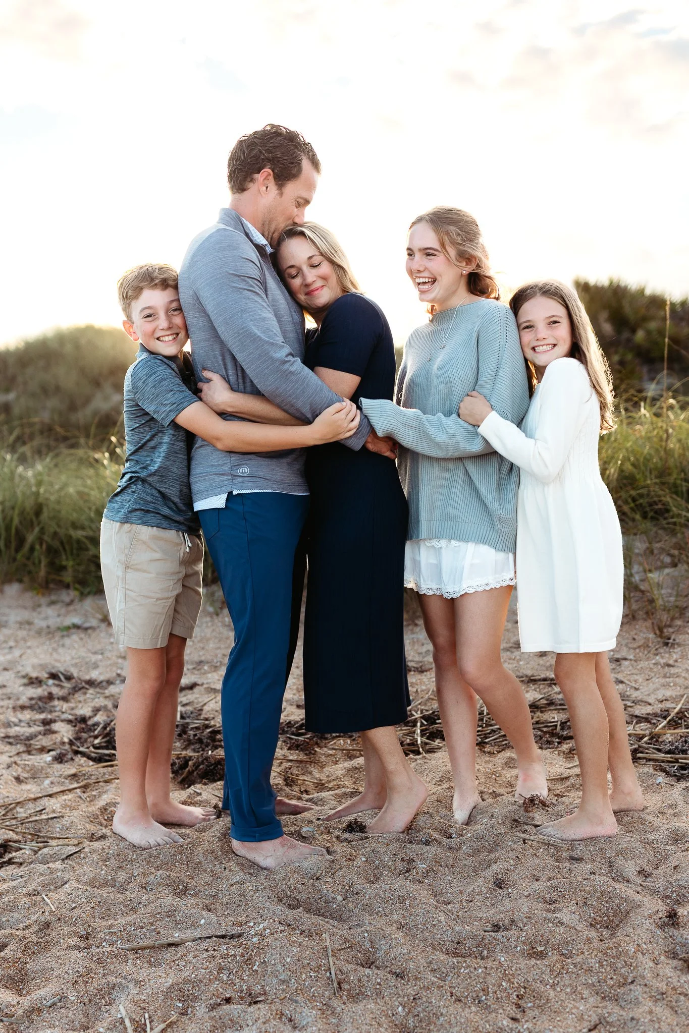 family standing on gunan beach dune and they are embraced the kids are laughing and the dad is kissing the mom whose eyes are closed