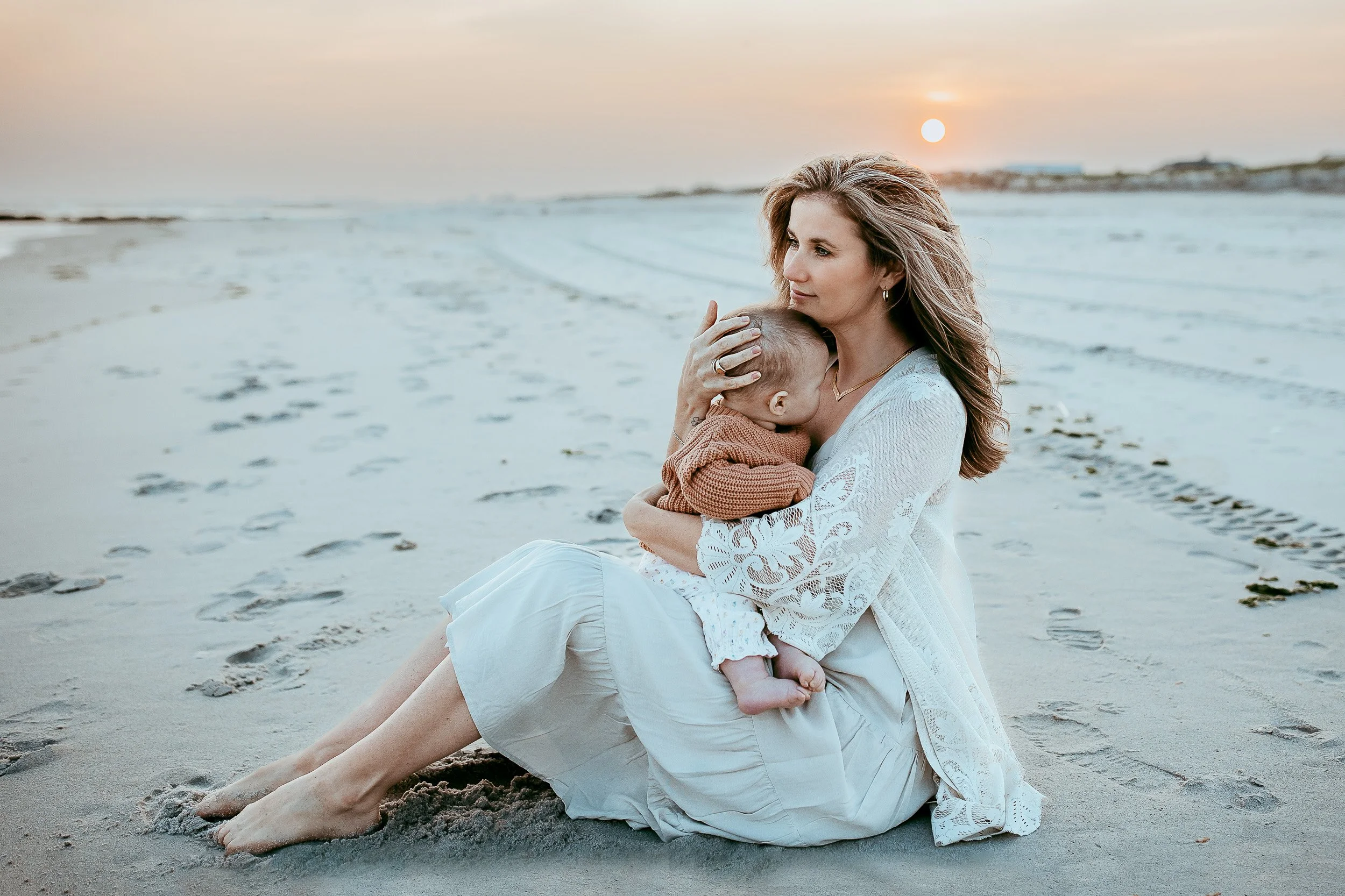 mother seated on the sand of the beach at sunset embracing her baby girl in a rust sweater and mom is wearing a lace kimono