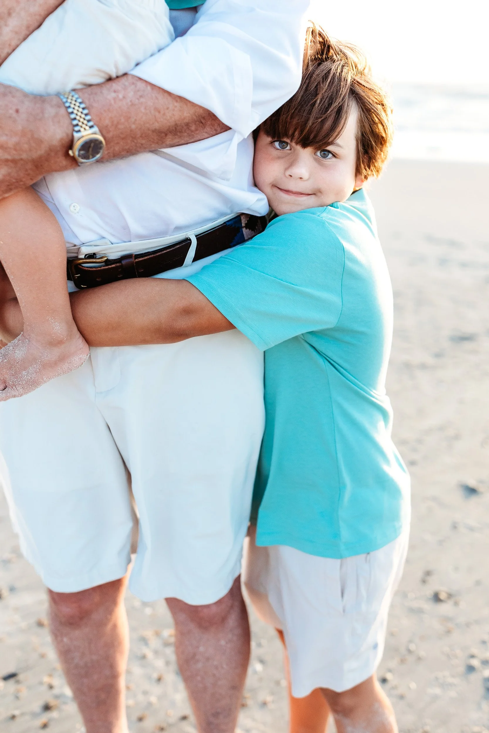 little boy in teal hugging his grandpa's leg during family photos on th beach