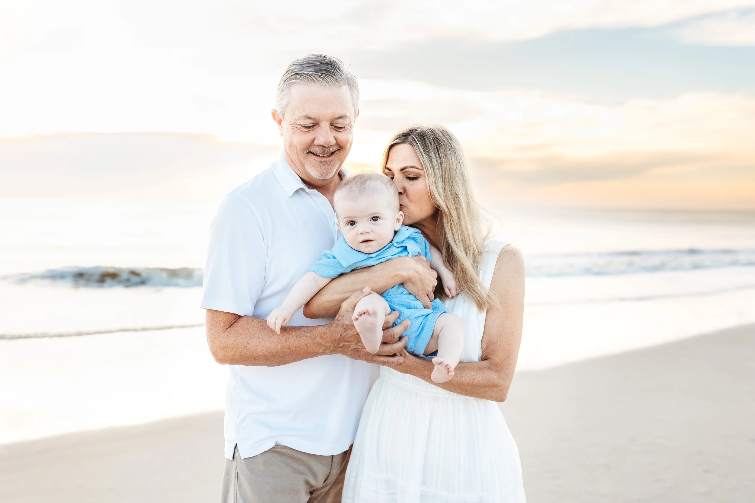 grandparents holding and kissing a baby on the beach at sunrise