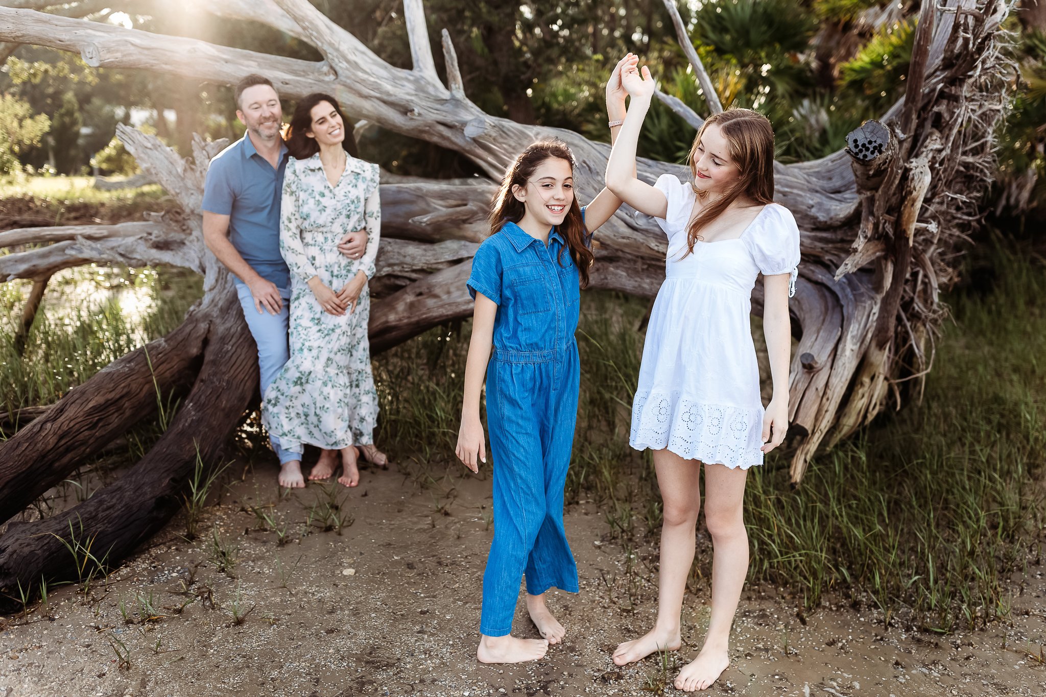 sisters dancing while parents watch in front of a driftwood tree