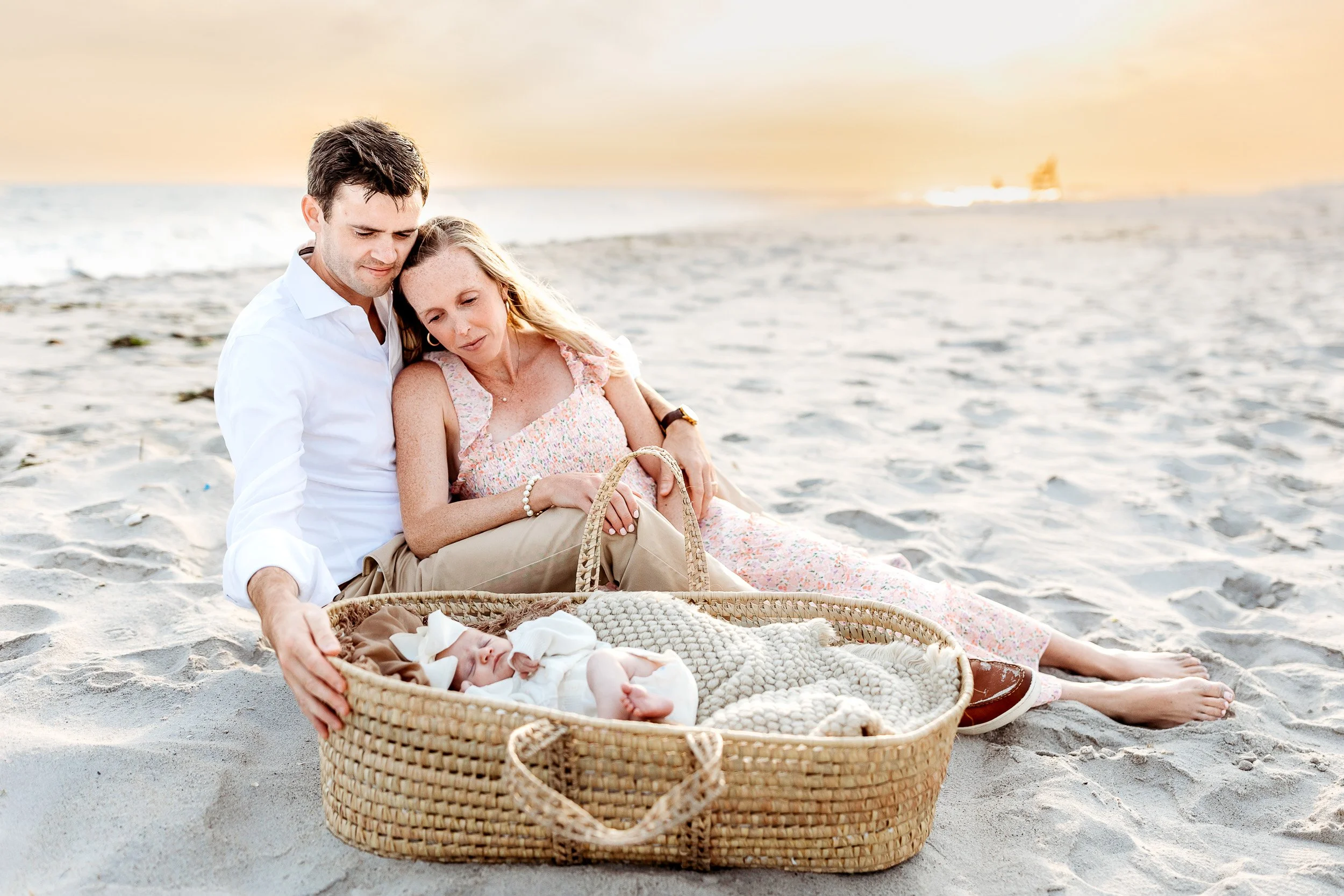 family on the beach on florida at sunset and a baby is in a Moses basket sleeping while the couple looks at her 
