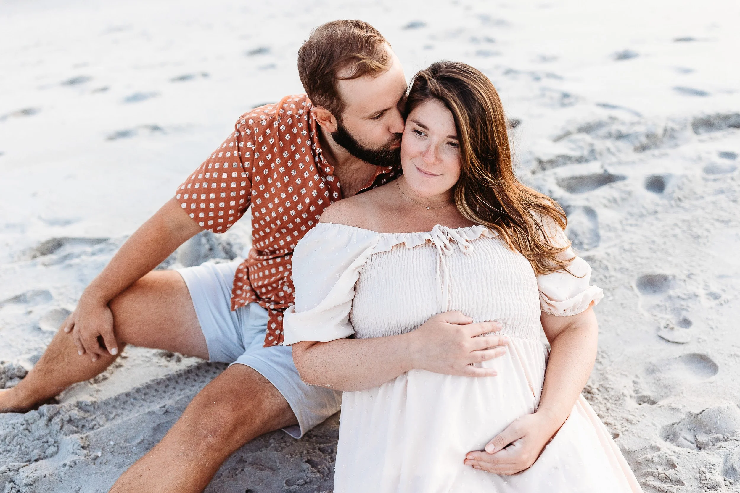 couple laying on the sand at the beach and pregnant woman touching her belly while husband kisses her