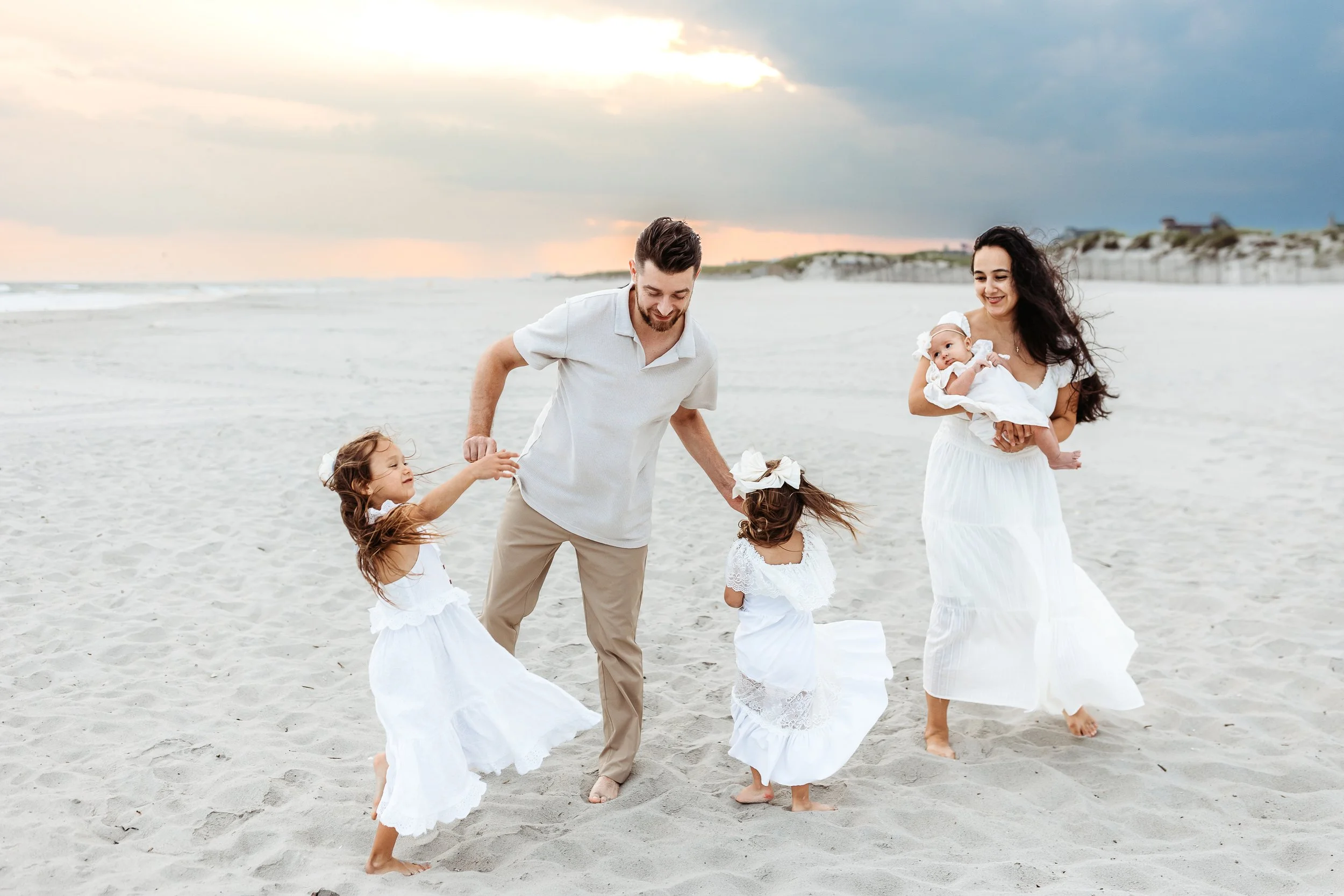 dad playing with toddler girls on the beach while mom watches and holds baby