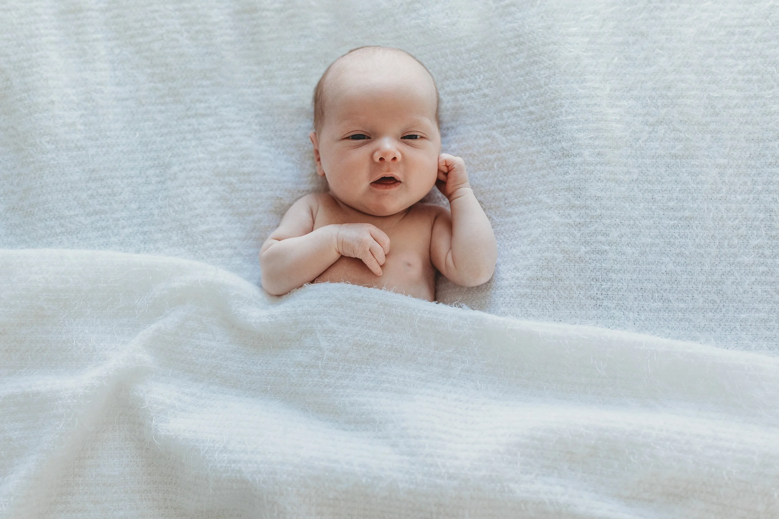baby on a white bed with a white blanket and his eyes are wide open and he is looking at the camera