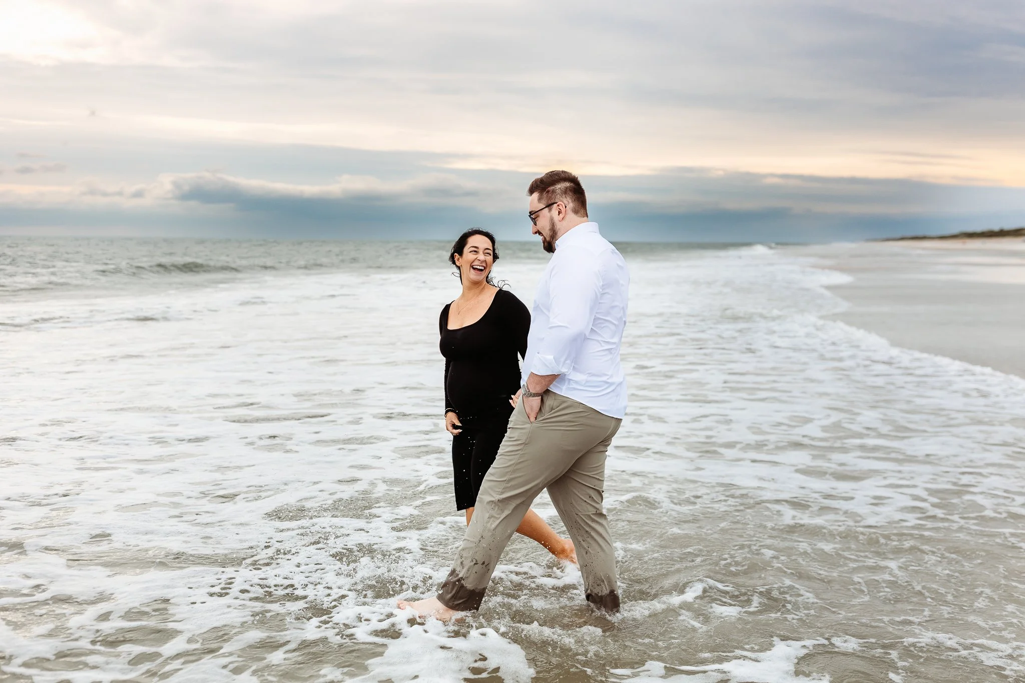 couple walking in ocean laughing and kicking waves and woman is pregnant and wearing black dress