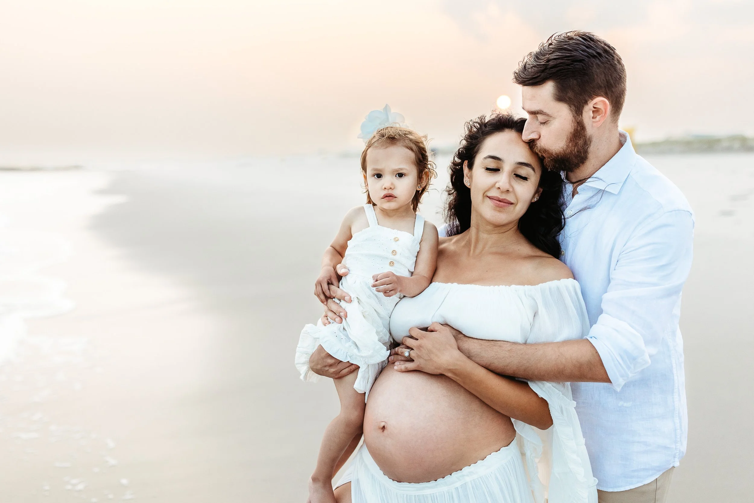 man kissing pregnant woman on the head and her belly is exposed and it is sunset
