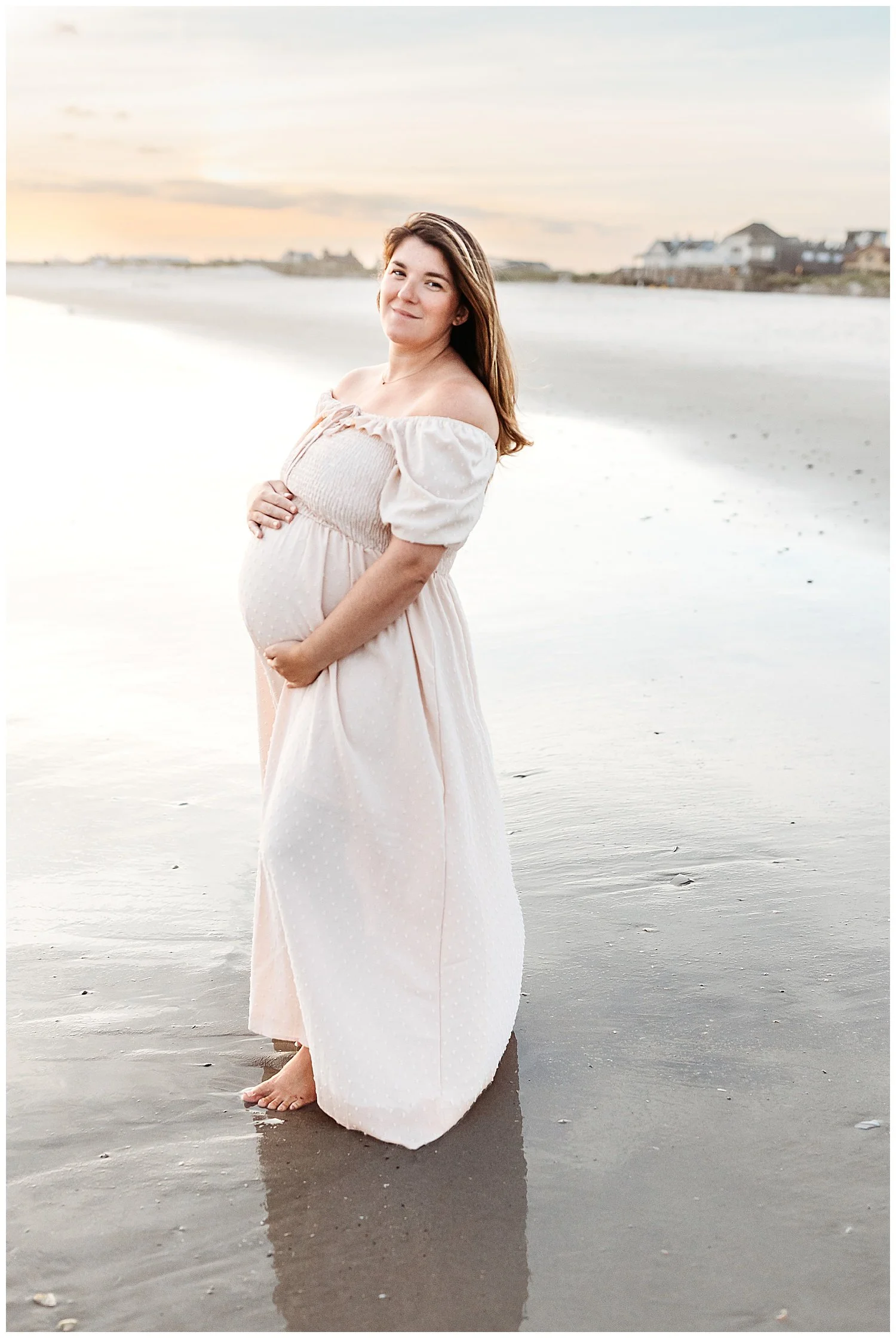woman on th beach for maternity portraits and she is holding her belly and smiling at the camera