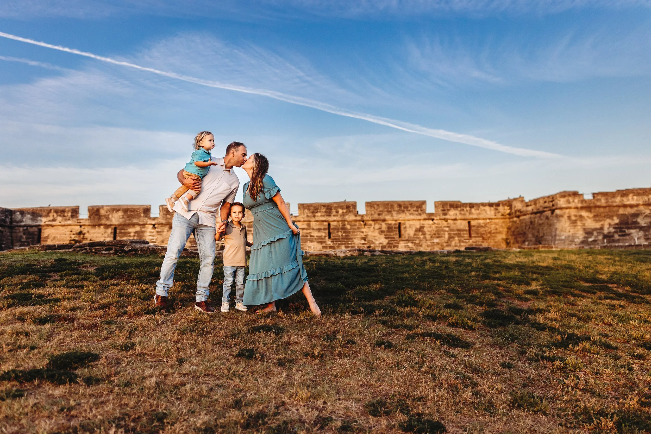 A family of four standing on grass near a stone fortress wall, with a clear blue sky and contrails above, as they share a kiss.