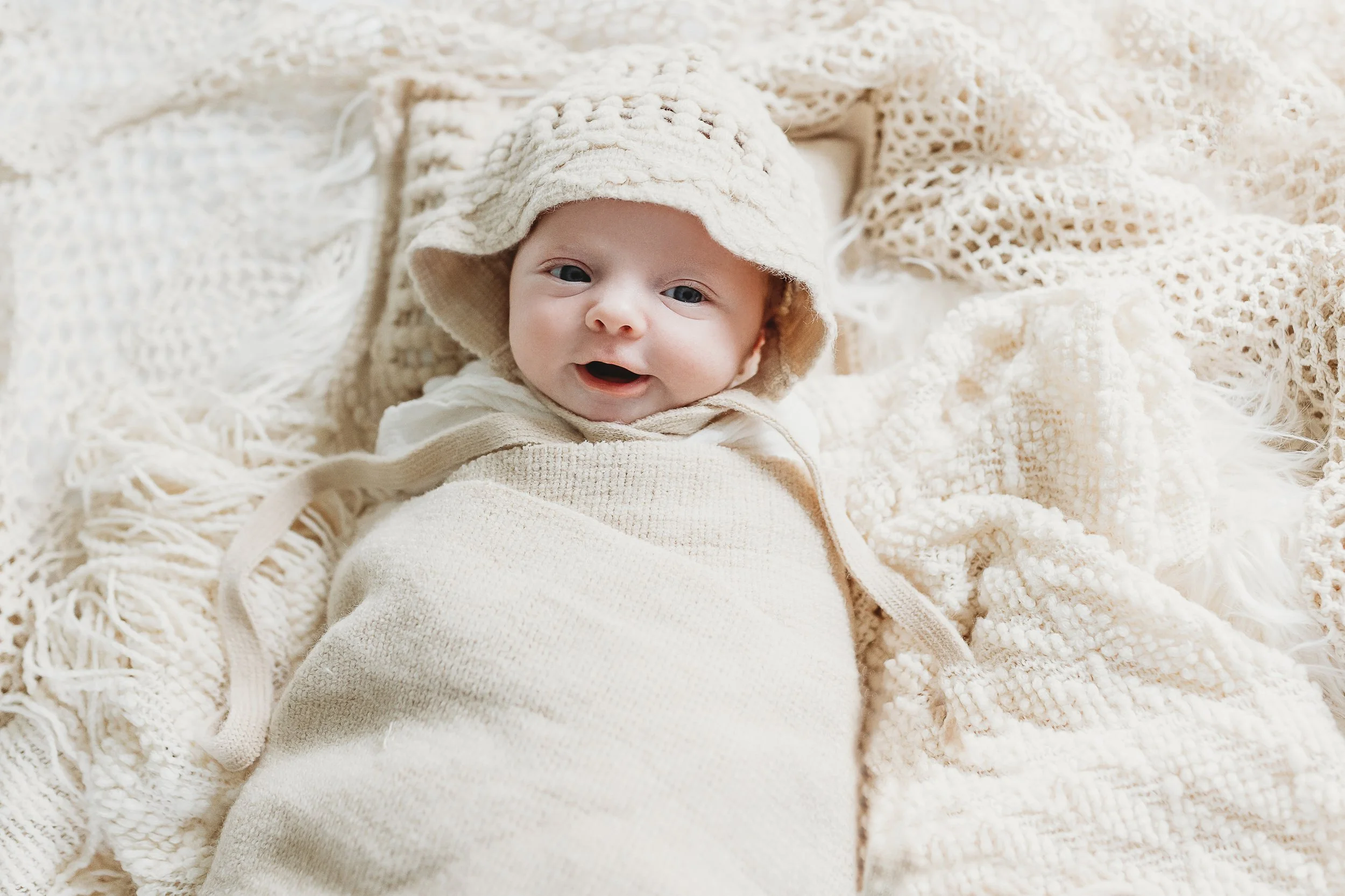 baby in cream colored bonnet and hat laying on boho crochet blanket and smiling during a newborn session in her home in st Augustine
