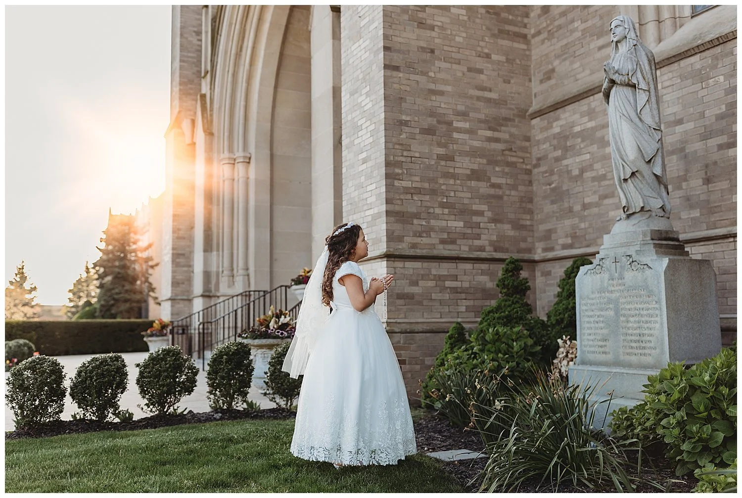 girl in white communion dress and veil praying in front of a Mary statue at a church and it is sunset