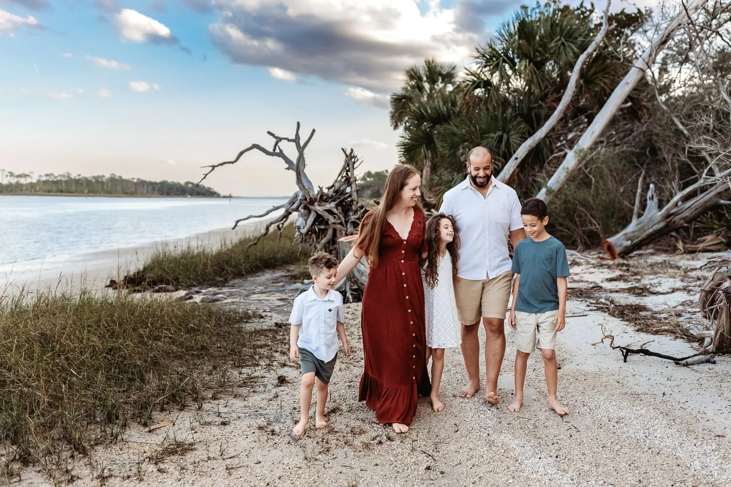 family in teal and rust wardrobe walking down a beach laughing with driftwood trees and intracoastal behind them