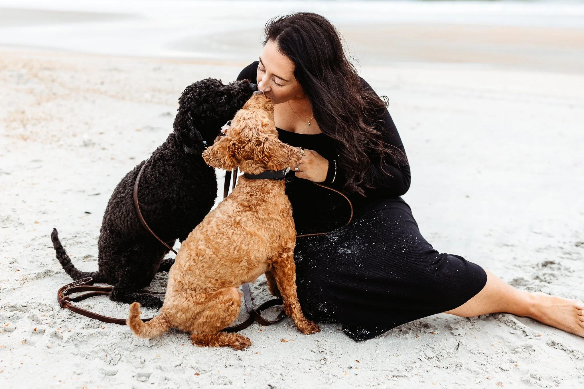 pregnant woman seated on sand kissing dogs