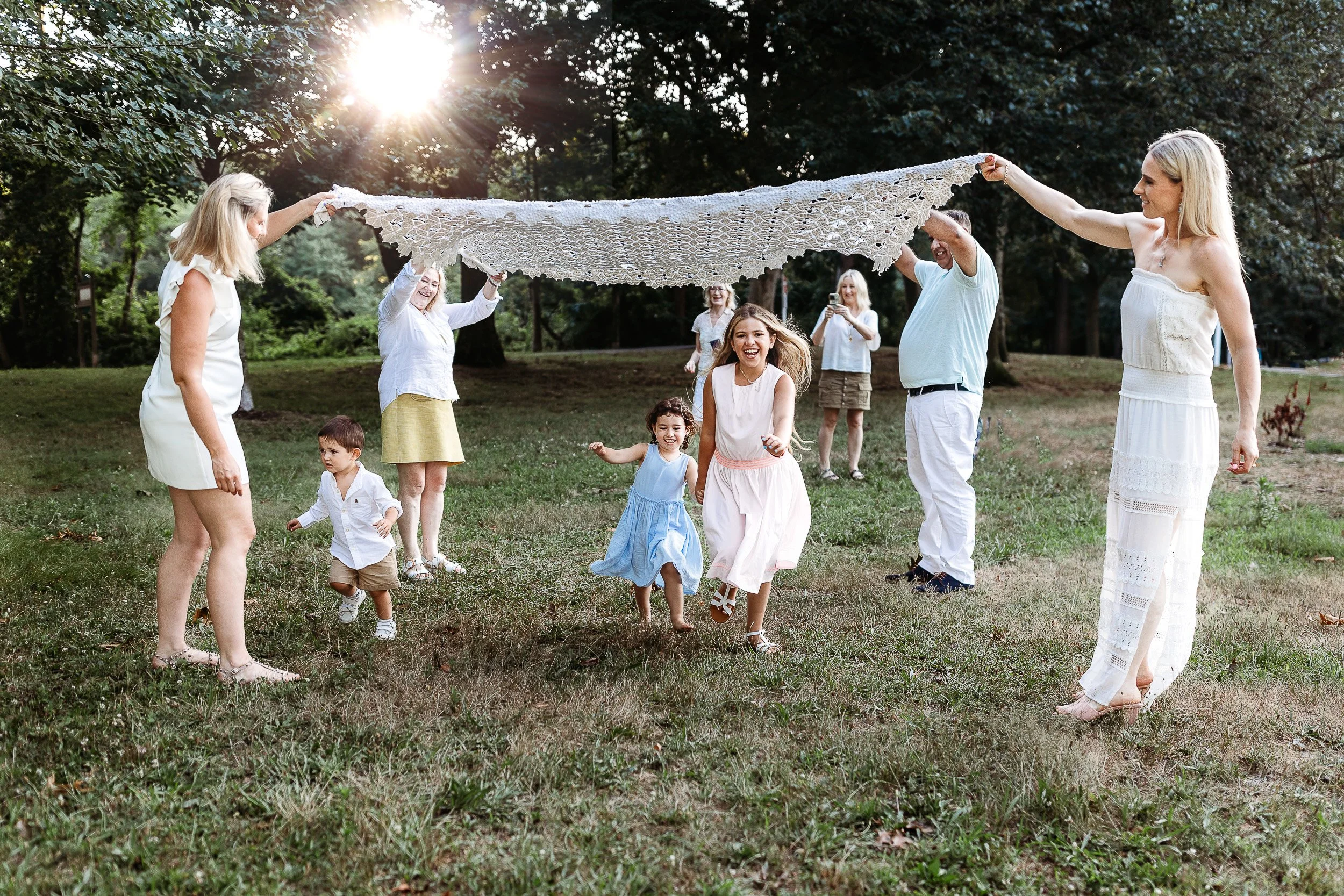 adult sisters playing popcorn with a white crochet blanket and toddler girls are running beneath the blanket and laughing.  Grand,a is taking a photo in the distance 