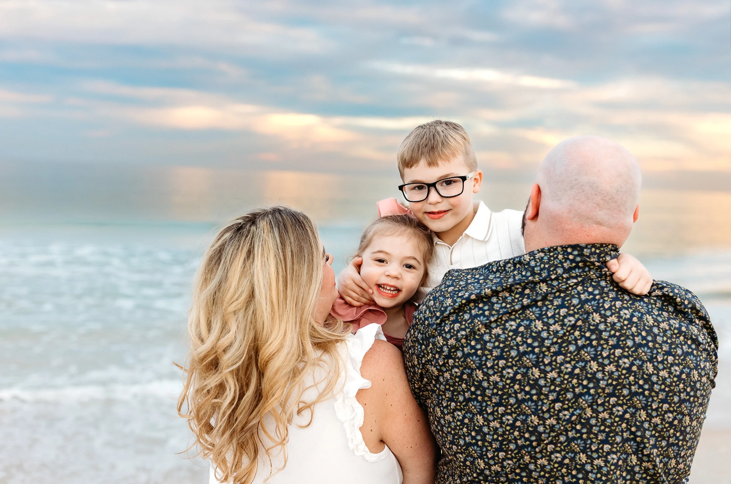 kids looking over parents shoulders and the ocean is behind them with a pink golden sunset sky