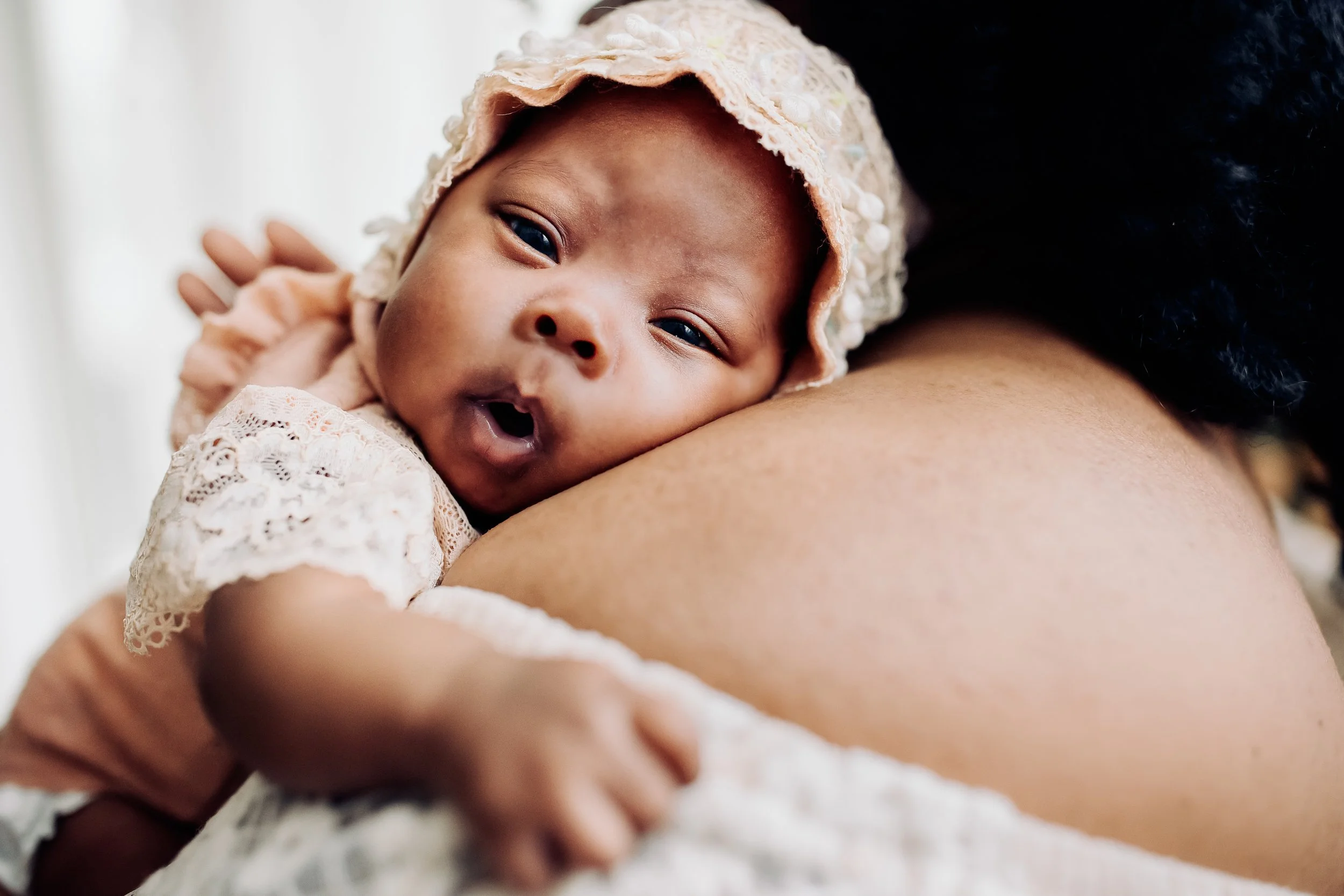 Close-up of a baby resting on an adult's shoulder, wearing a lace bonnet and outfit, with the baby’s mouth open and eyes slightly squinted.