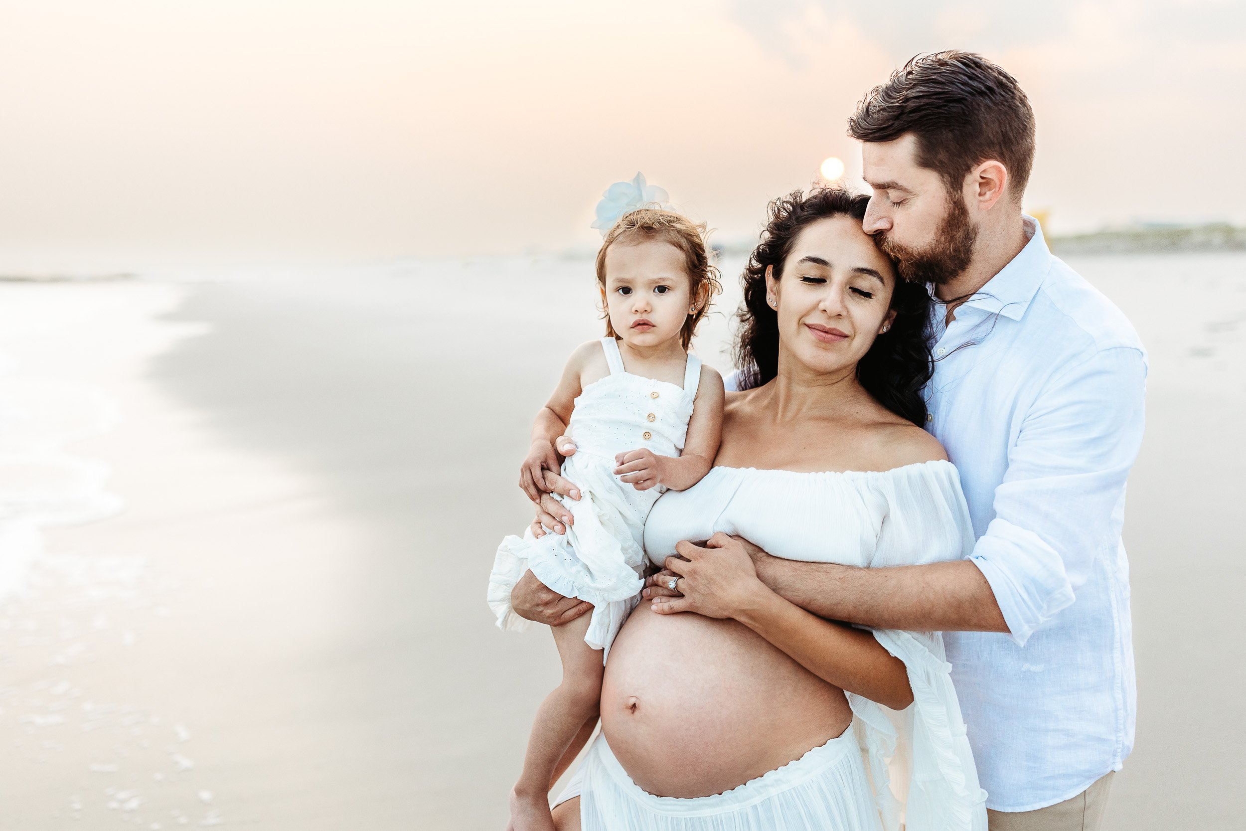 husband kissing wife on the beach at sunset and her eyes are closed and they are both holding her pregnant belly