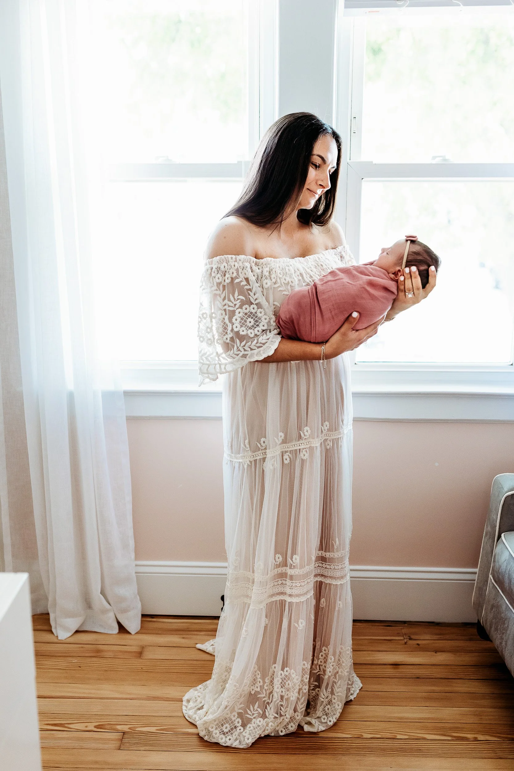 woman in ivory lace off the shoulder gown holding a baby in a pink swaddle and she is in front of a window and smiling