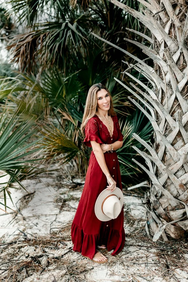 Woman in a red maxi dress standing on sandy ground next to palm trees, holding a hat, in a tropical setting.