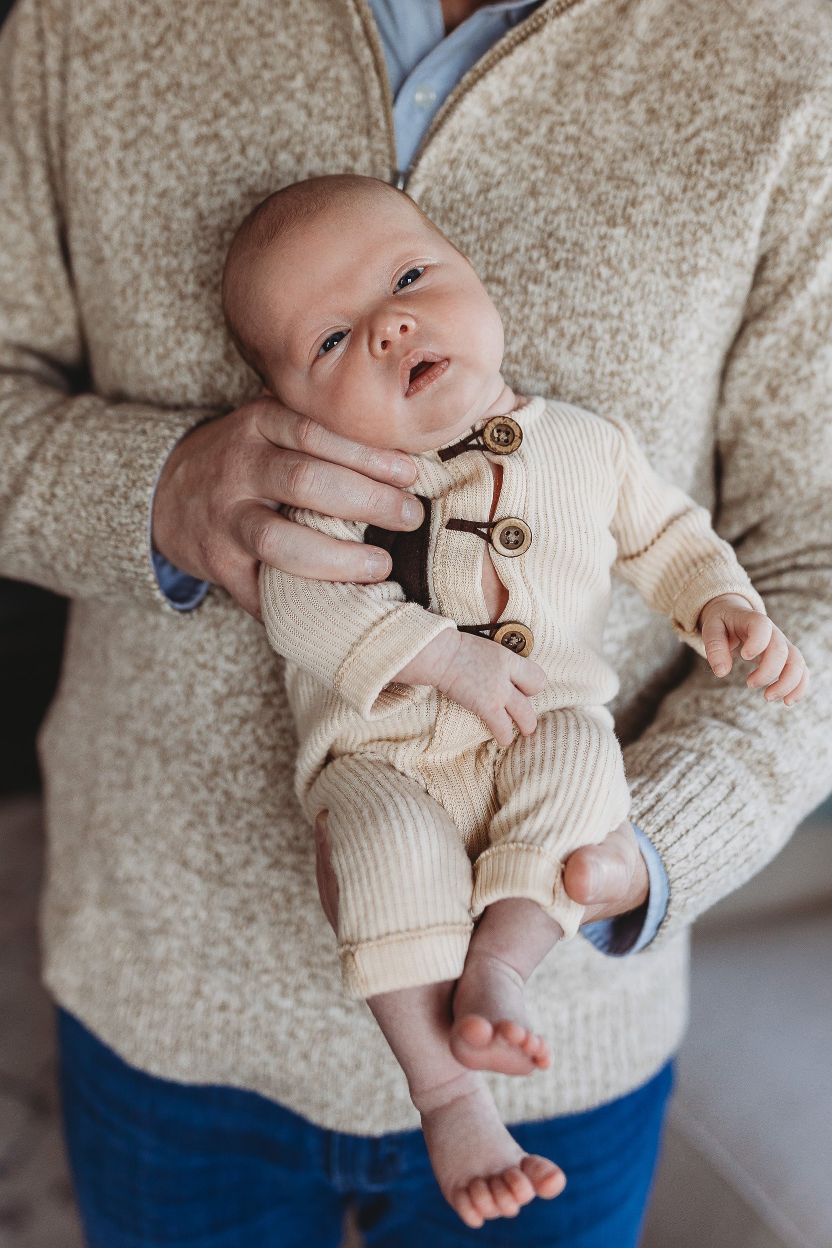 close up of a baby in his dad's hands wearing tan knit vintage style onesie and his eyes are sleepy and they are in their home for newborn photos in the Ponte Vedra florida area