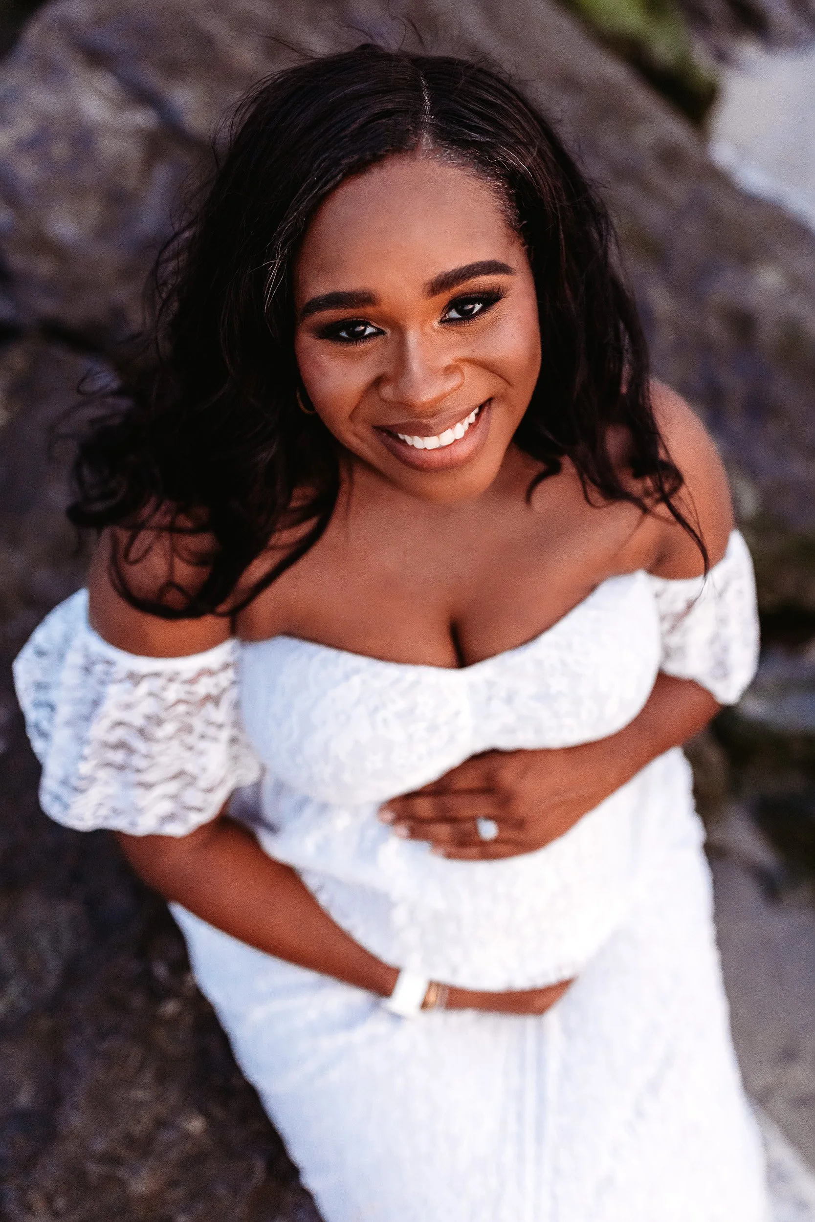 close up portrait of a beautiful woman pregnant and holding belly wearing white lace gown and looking up at camera during a maternity photo session on st augustine beach