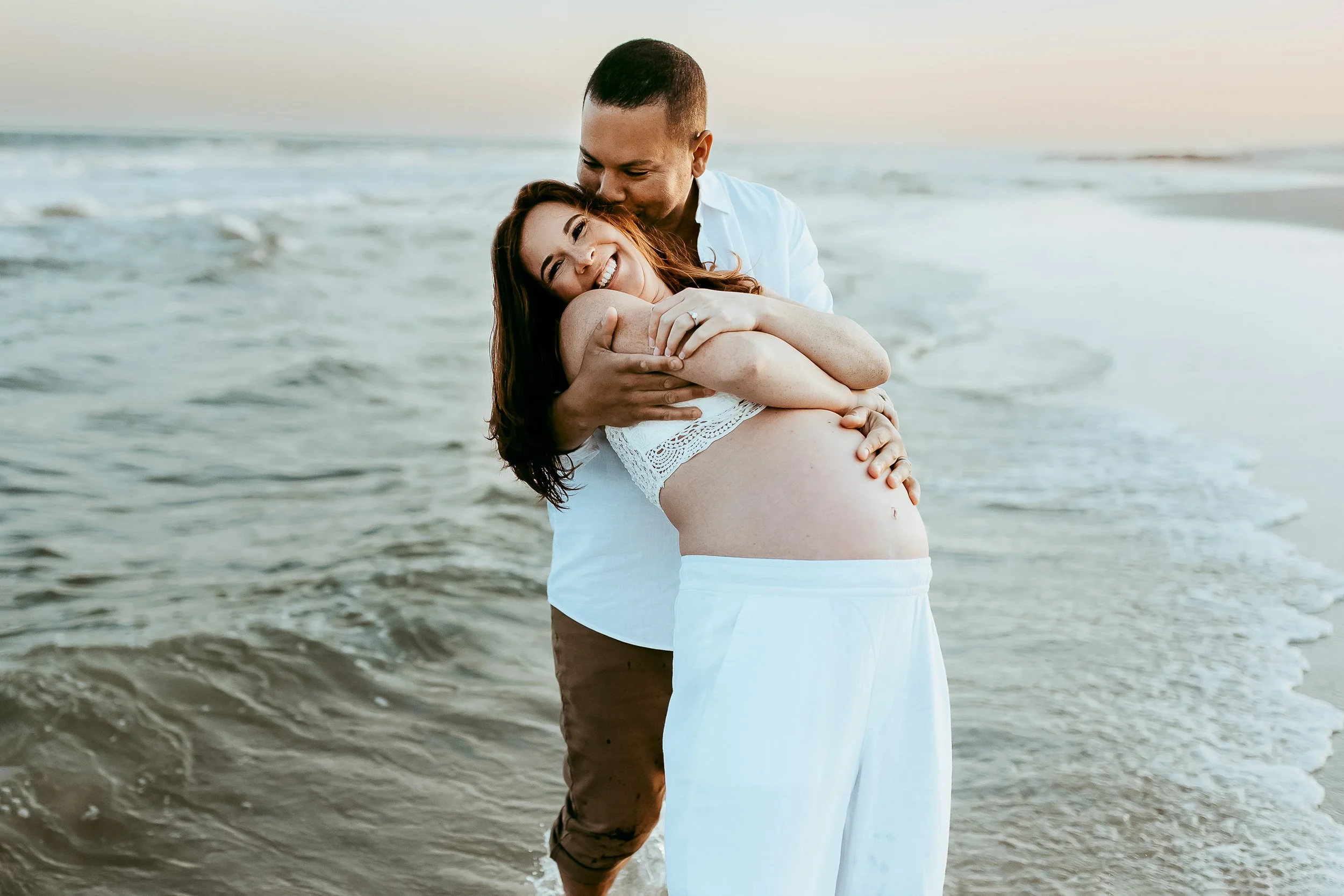 woman being leaned back by husband and he is kissing her ear and she is laughing they are on a beach in st Augustine for maternity couples photos