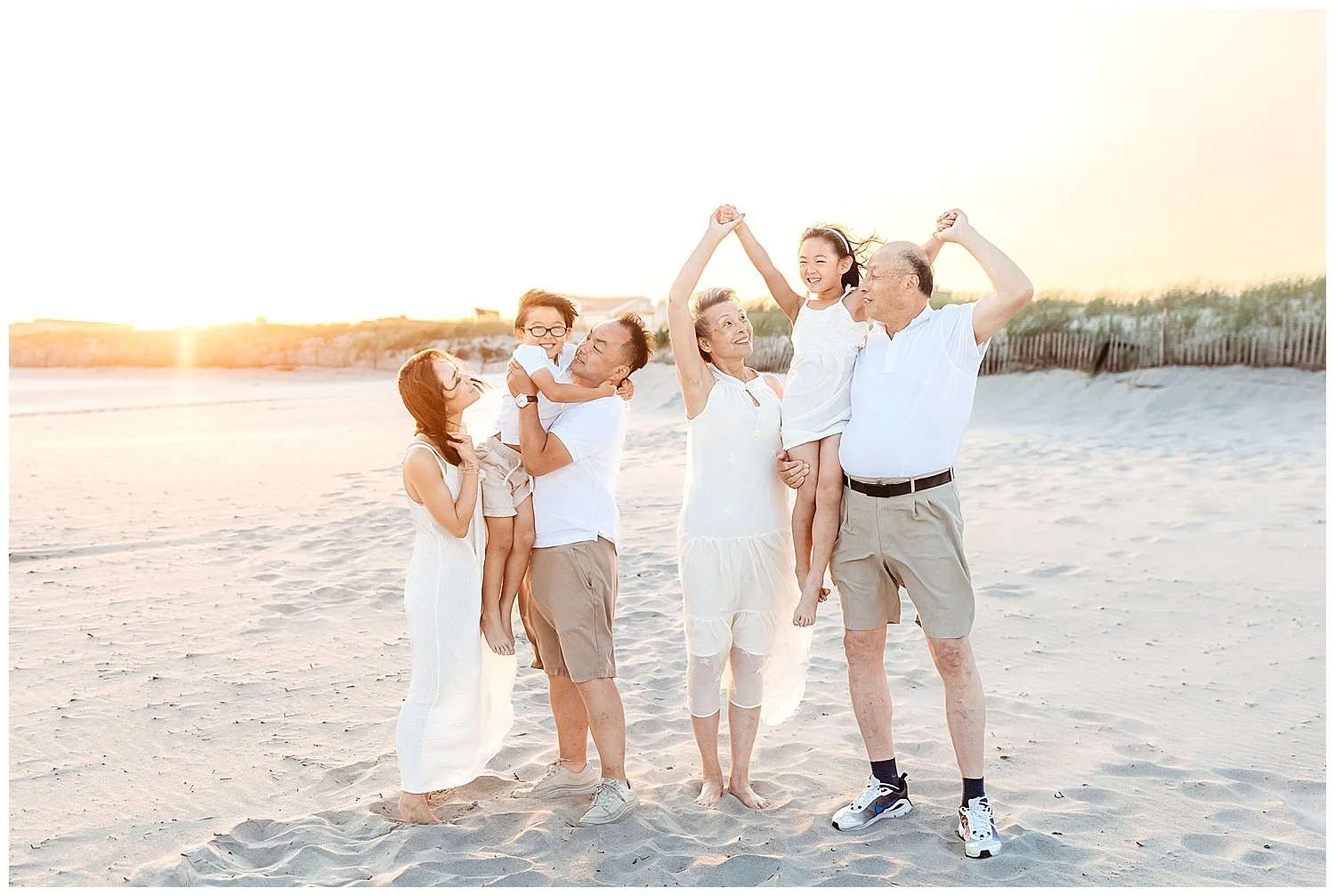 girl on grandpa's shoulders holding grandma's hands boy being held by dad on beach at sunset all laughing