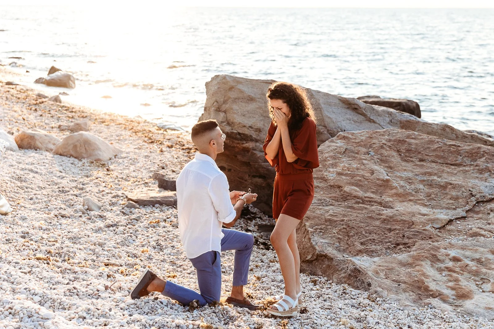 A man kneeling with a ring in his hand before a woman on a rocky beach, both are emotional and smiling.